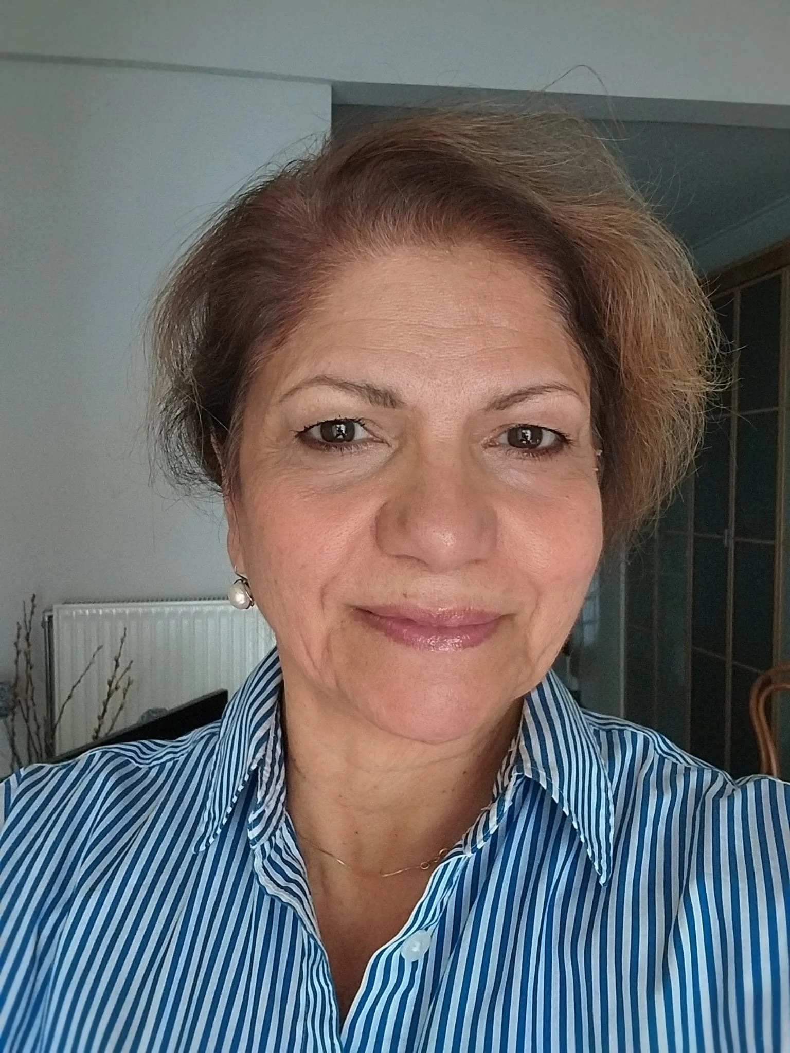 Close-up of a woman with short, wavy, light brown hair wearing a blue and white striped shirt, standing in a home interior.
