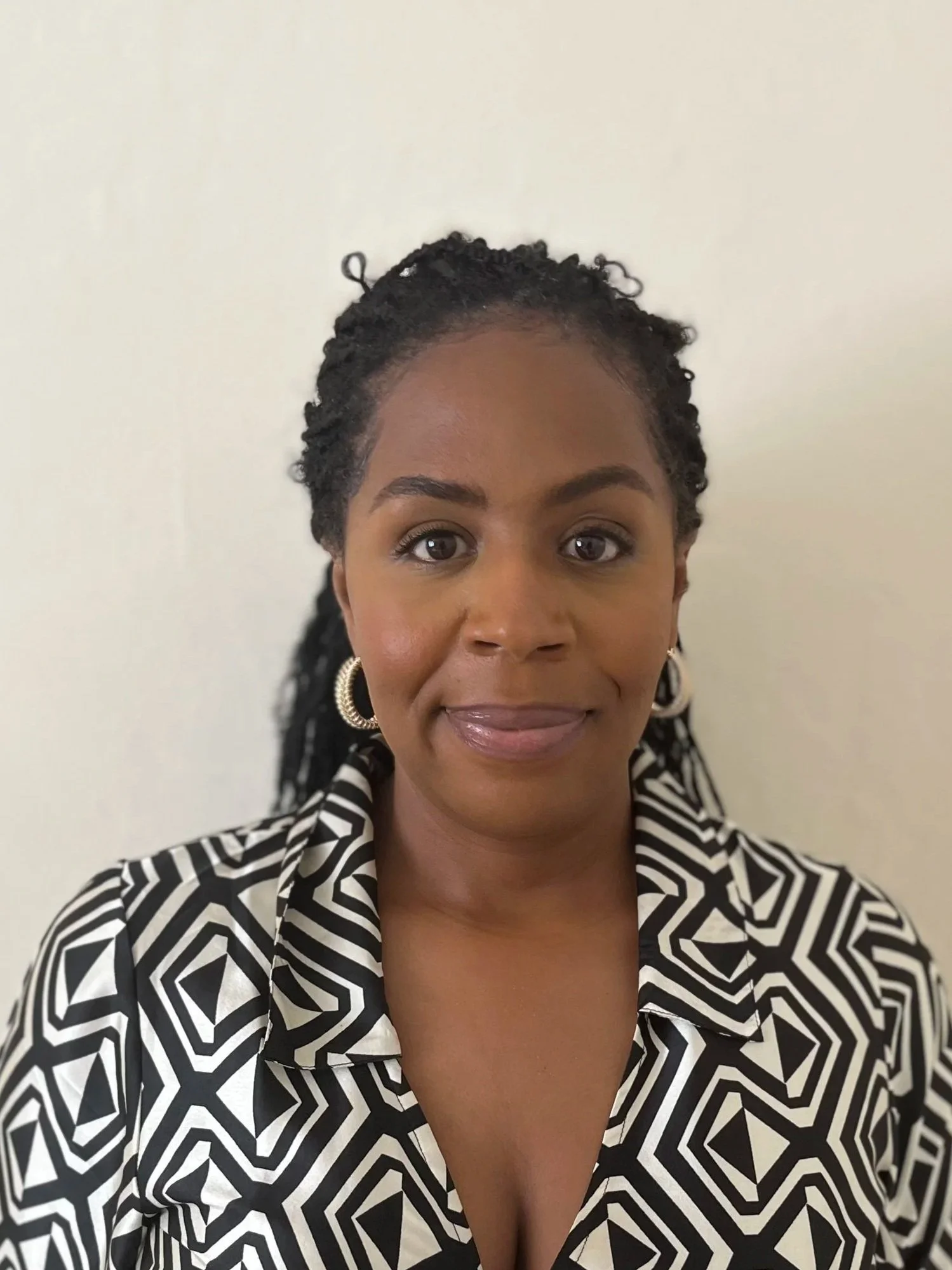Close-up of a woman with dark braided hair, wearing hoop earrings and a black and white geometric patterned blazer, standing against a plain light-colored background.