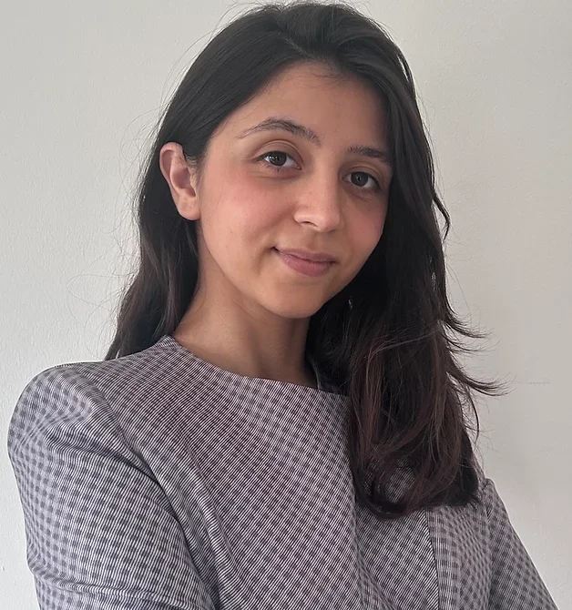 A young woman with long dark hair and light skin, wearing a checkered blouse, standing against a plain white background.