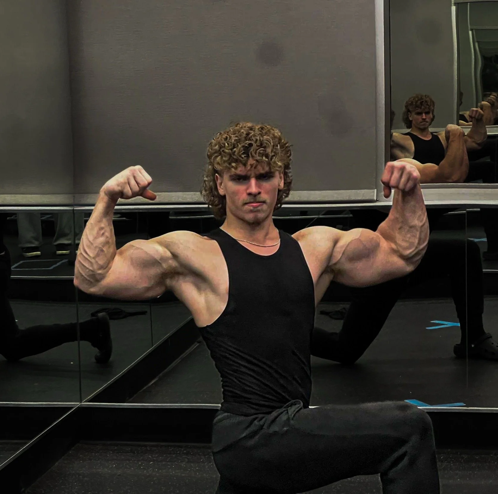 A young man with curly hair in a black tank top and black pants flexes his biceps in front of a mirror in a gym, showing muscular arms.