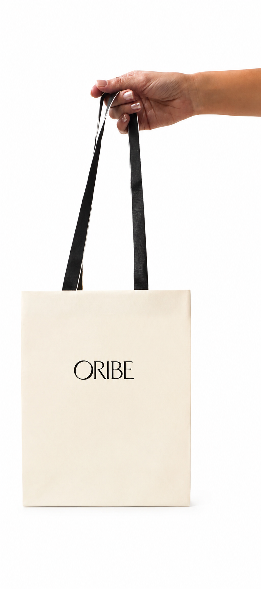 A hand holding black-and-white satin handles of a beige shopping bag with the brand name 'ORIBE' printed on it, against a white background.