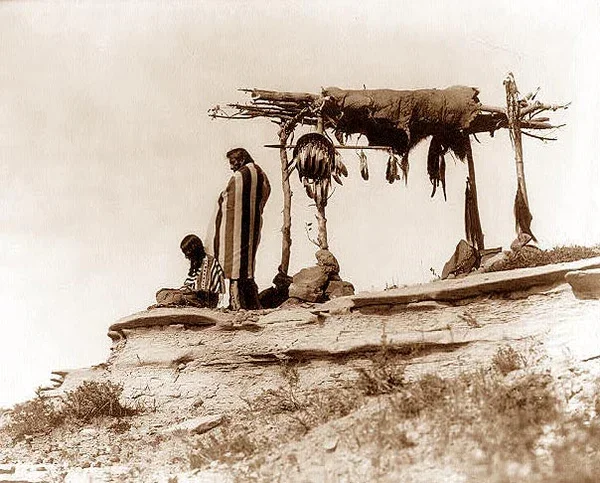 A sepia colored photo that depicts two native americans preparing for a sky burial