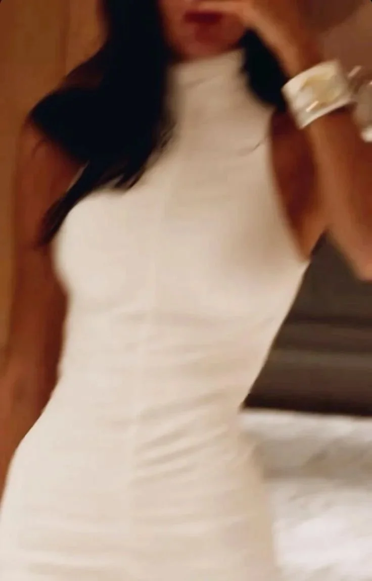 A woman with long, dark hair wearing a tight, sleeveless white dress holding a small cup or container, possibly in a casual or social setting.