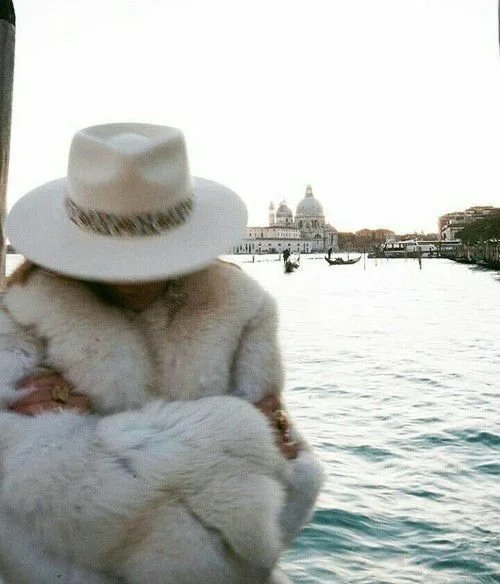 Person wearing a white hat and a fur coat stands near water with gondolas and classic European architecture in the background.