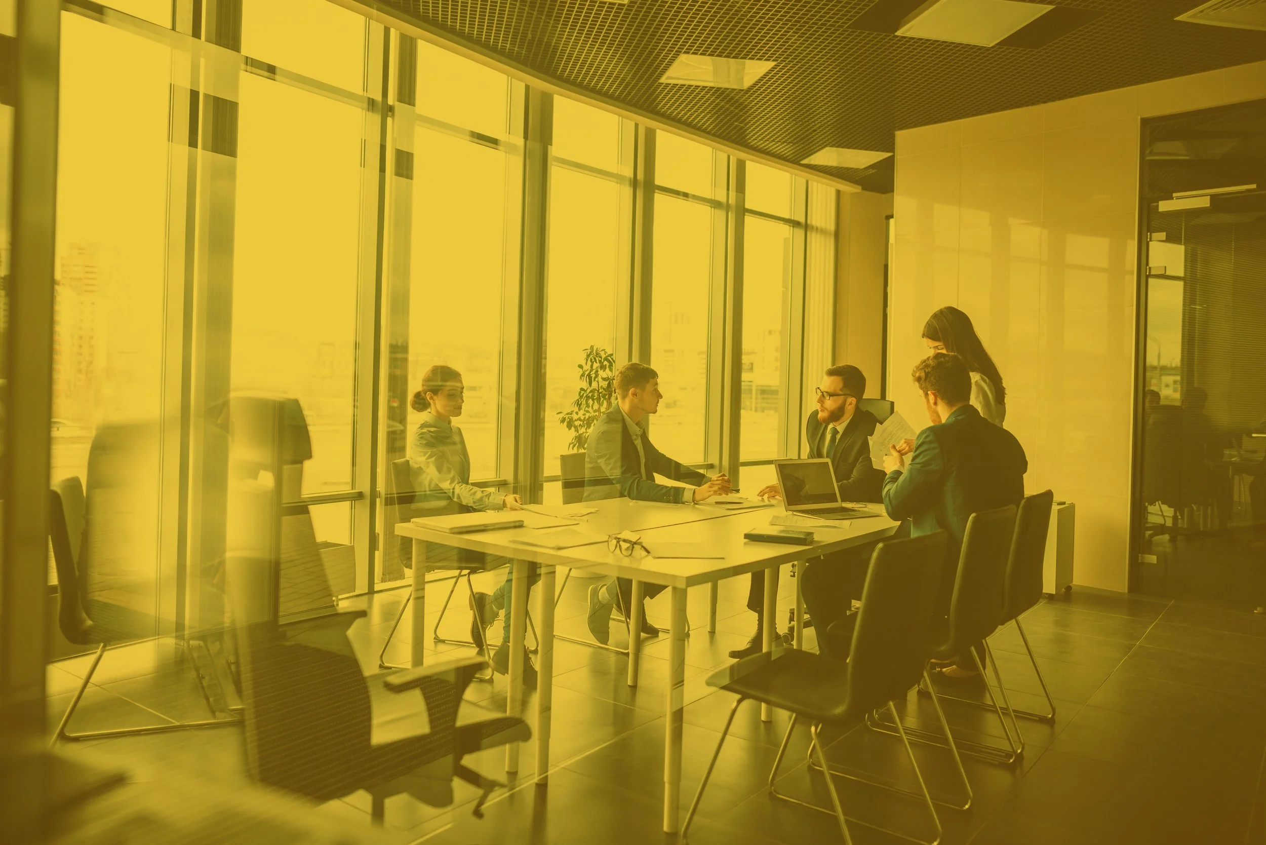 Business meeting in a modern conference room with large glass windows, five people sitting around a table, two women and three men, discussing and taking notes, some using laptops and documents.