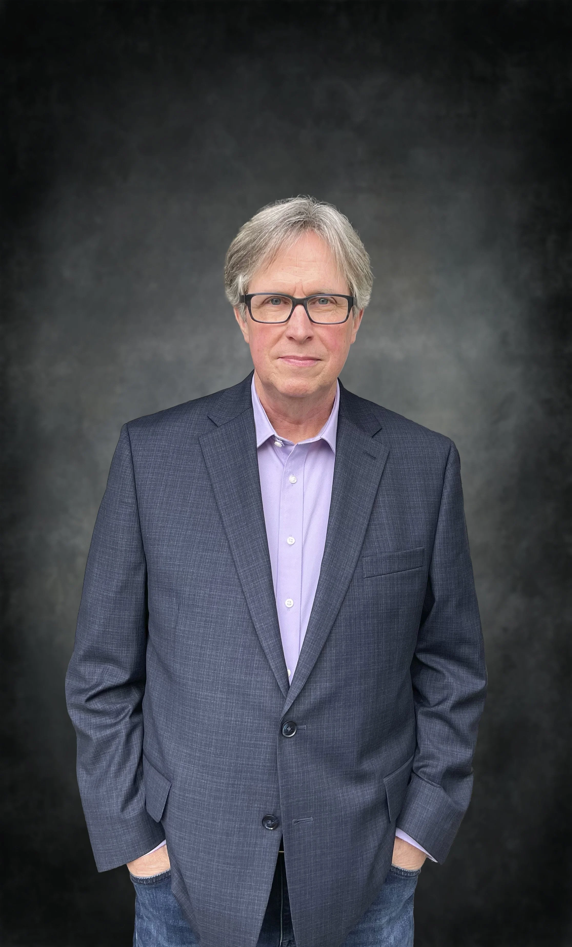 A middle-aged man with gray hair, wearing glasses, a dark gray suit jacket, a light purple dress shirt, and jeans, standing against a dark, textured background.