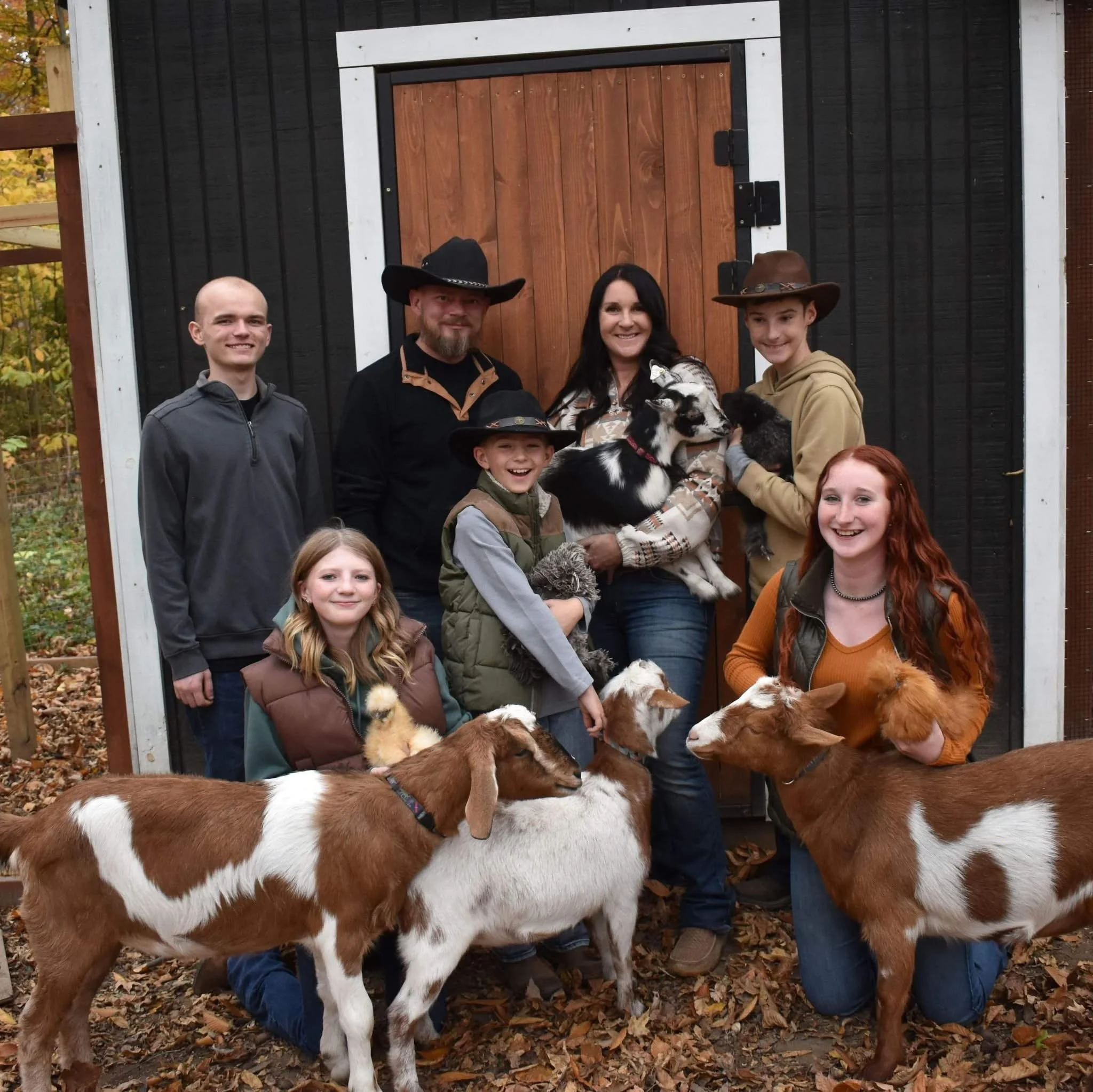 Family of seven posing outdoors in fall, with goats and puppies in front of a wooden barn door. Some family members wear cowboy hats, and everyone is smiling.