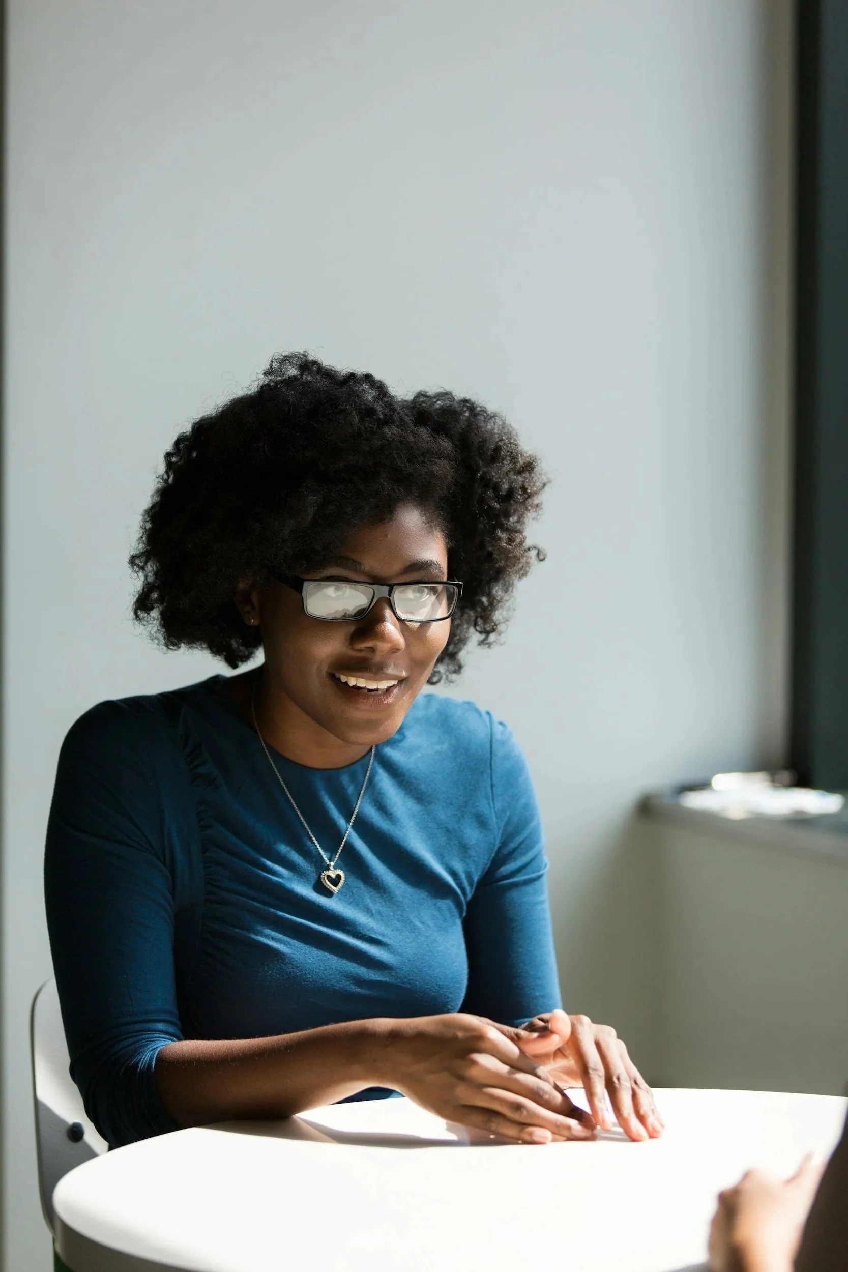 A smiling woman with glasses and curly hair wearing a blue long-sleeve shirt, sitting at a white round table and engaging in conversation.