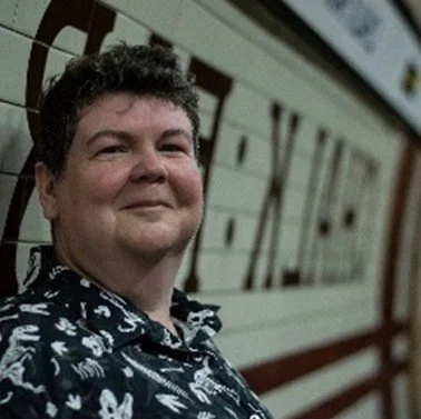 Dr Gillian Bush, Wellcare Health, Beverley smiling and wearing a black shirt with a white pattern, standing in front of a wall with large letters.
