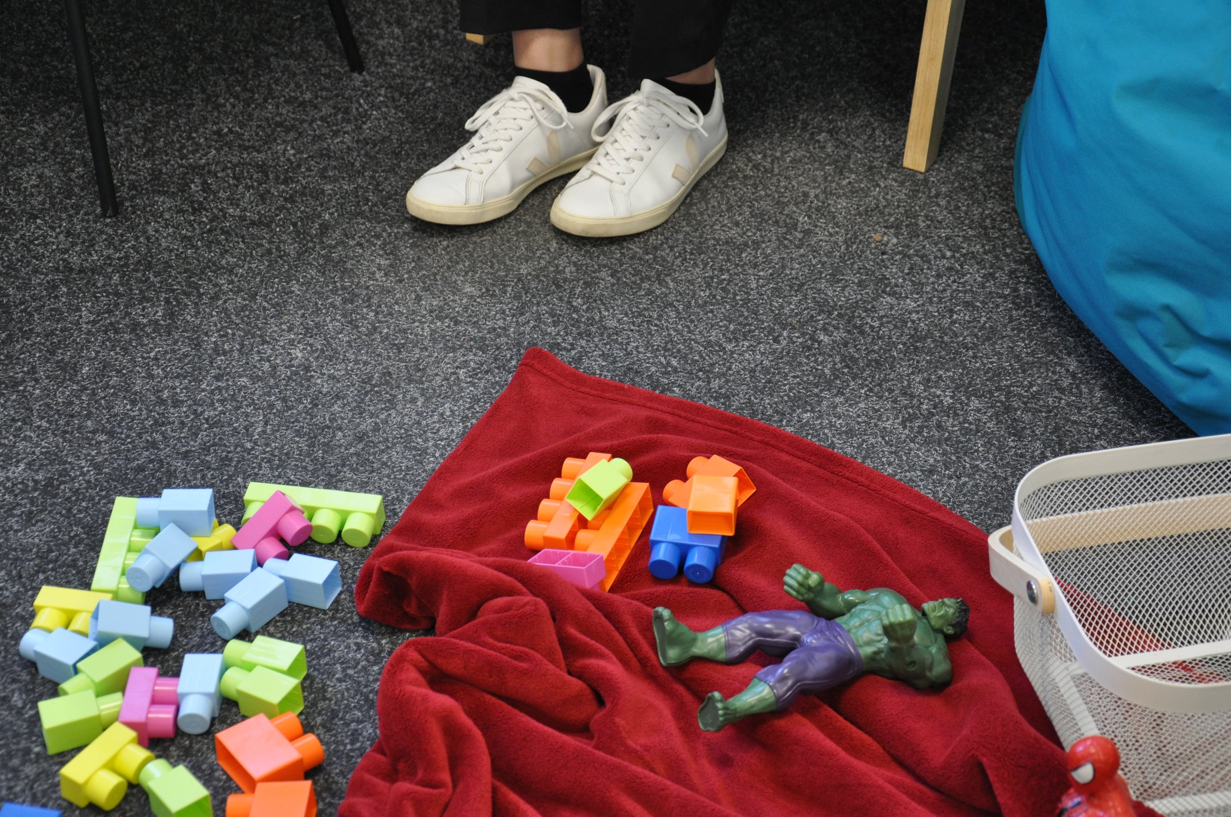 Colorful building blocks scattered on a gray carpet, with a red cloth, toy figures, and a white basket nearby. A person's feet in white sneakers and black socks are visible, sitting on a chair.