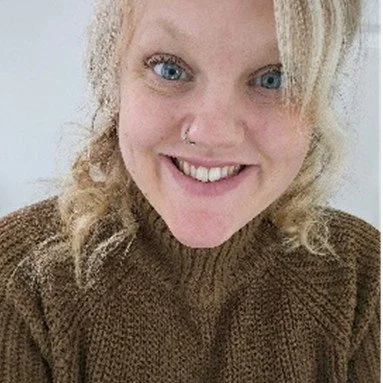 Emma Banks, Wellcare Health, Beverley with blonde curly hair, blue eyes, and a nose piercing smiling at the camera, wearing a brown sweater.