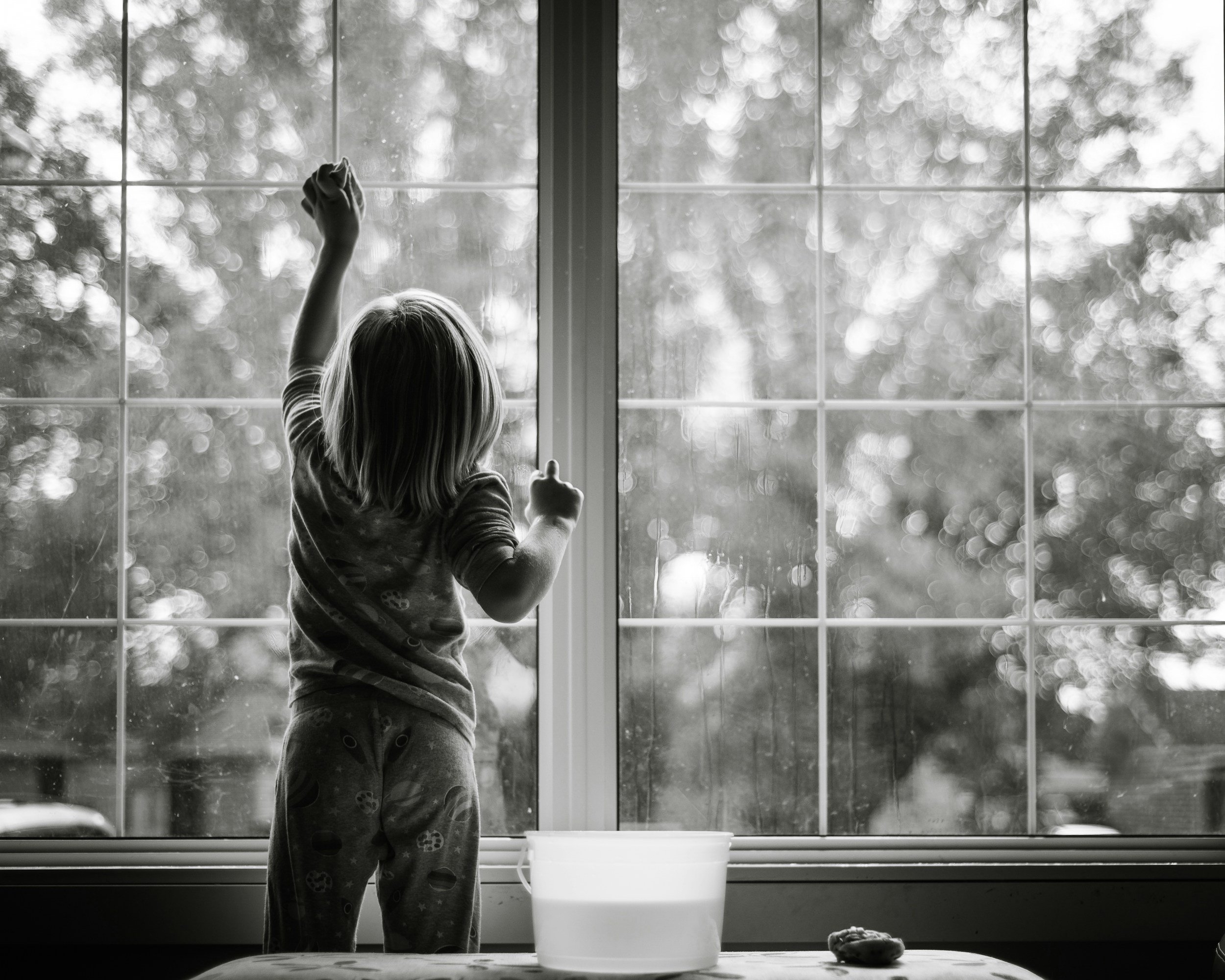A child looks out a window while cleaning it with a cloth. The window has multiple panes, and outside there are blurred trees and sky. The room has a table with a white bowl and a scrubbing sponge.