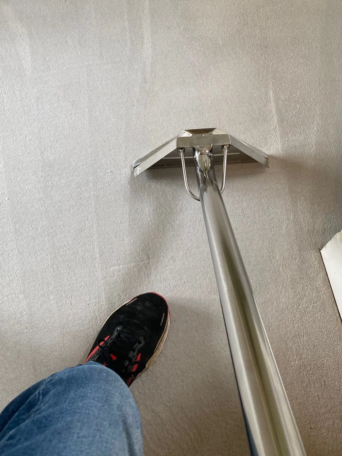 A person standing on a beige carpeted floor, wearing black sneakers with red accents and blue jeans, next to a metal mop or cleaning tool.