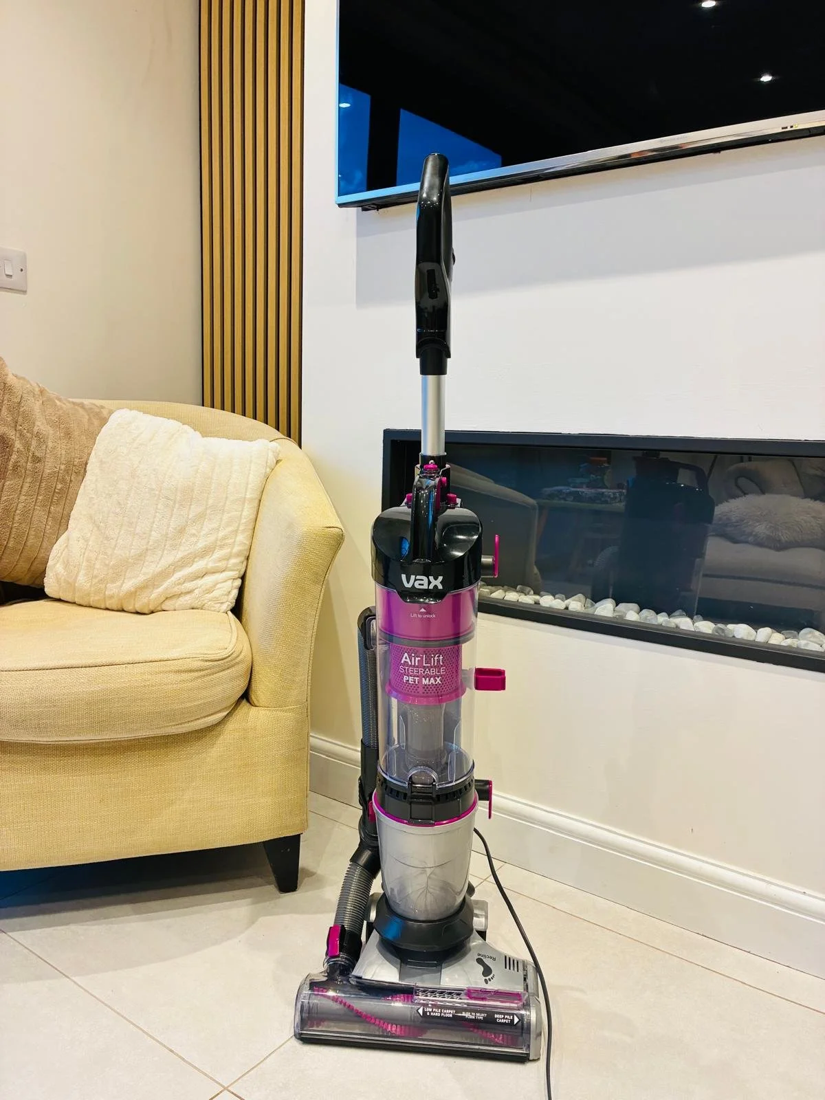 A pink and black Vax AirLift stearable vacuum cleaner standing on a light-colored tiled floor next to a beige sofa with pillows and a white wall with a vent. A flat-screen TV is mounted on the wall behind the vacuum.