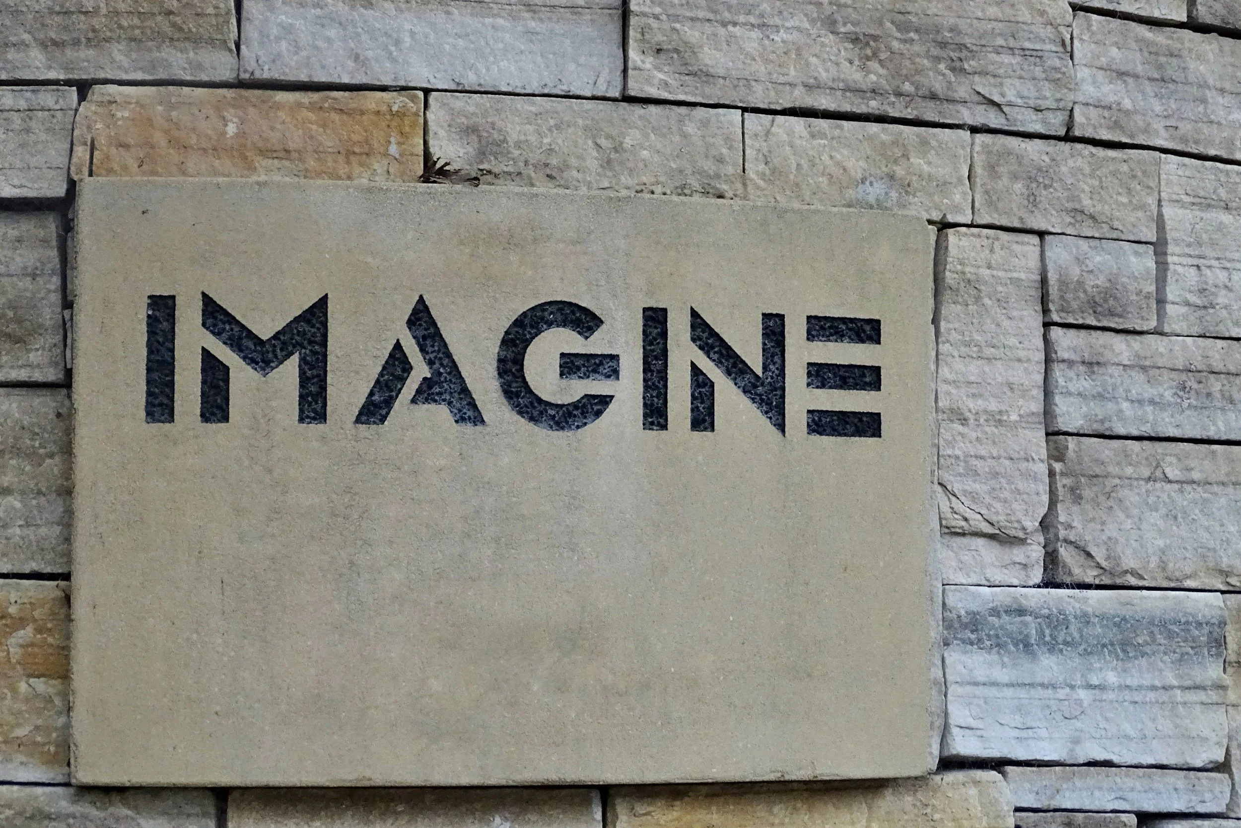 Panneau en pierre avec l'inscription 'IMAGINE' en lettres noires, fixé sur un mur en pierre