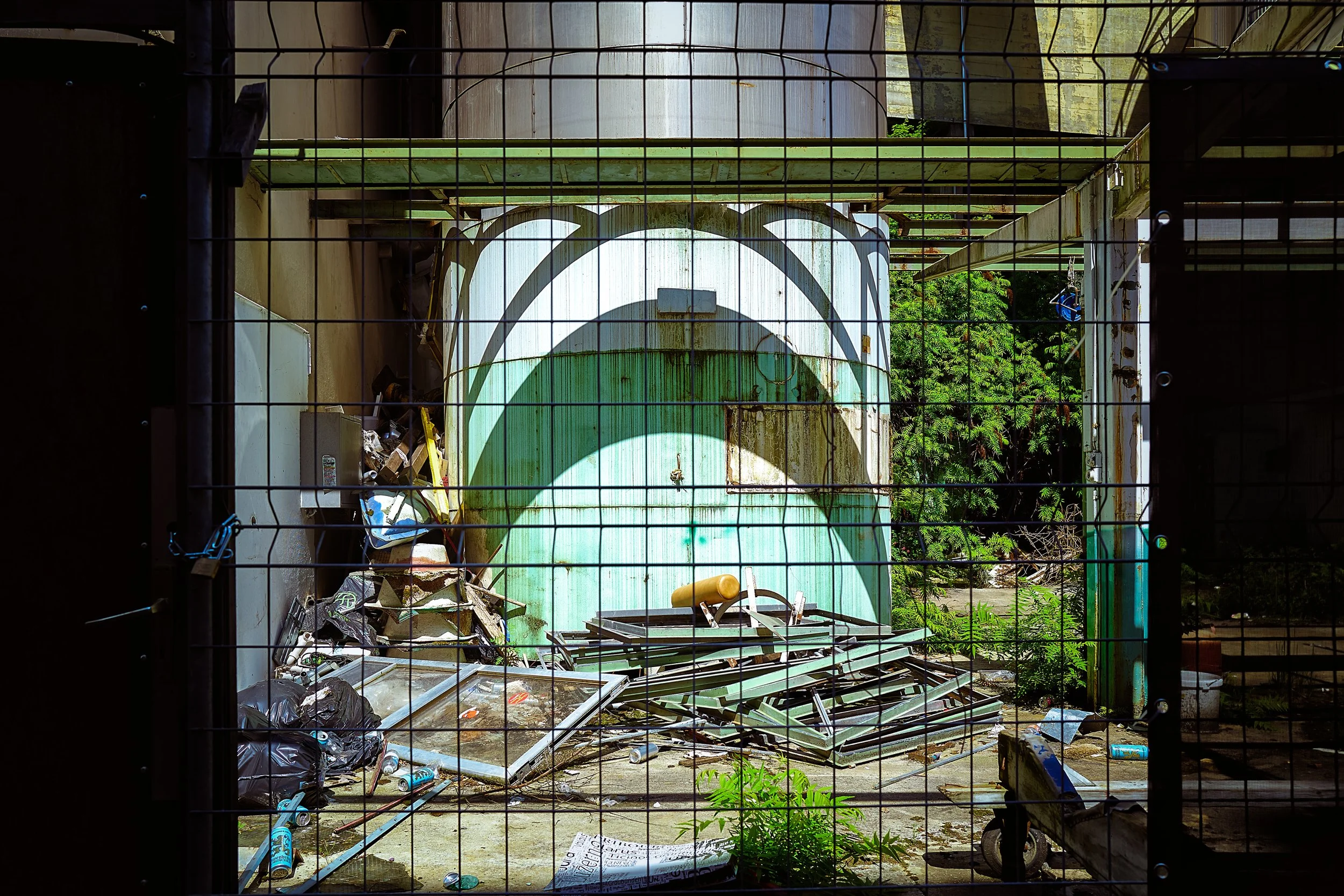 Un espace abandonné avec un grand trou rouge dégageant des objets métalliques et une structure de toiture verte sur fond de verdure. Sont visibles des déchets, des déchets en plastique, et un panneau en verre brisé.