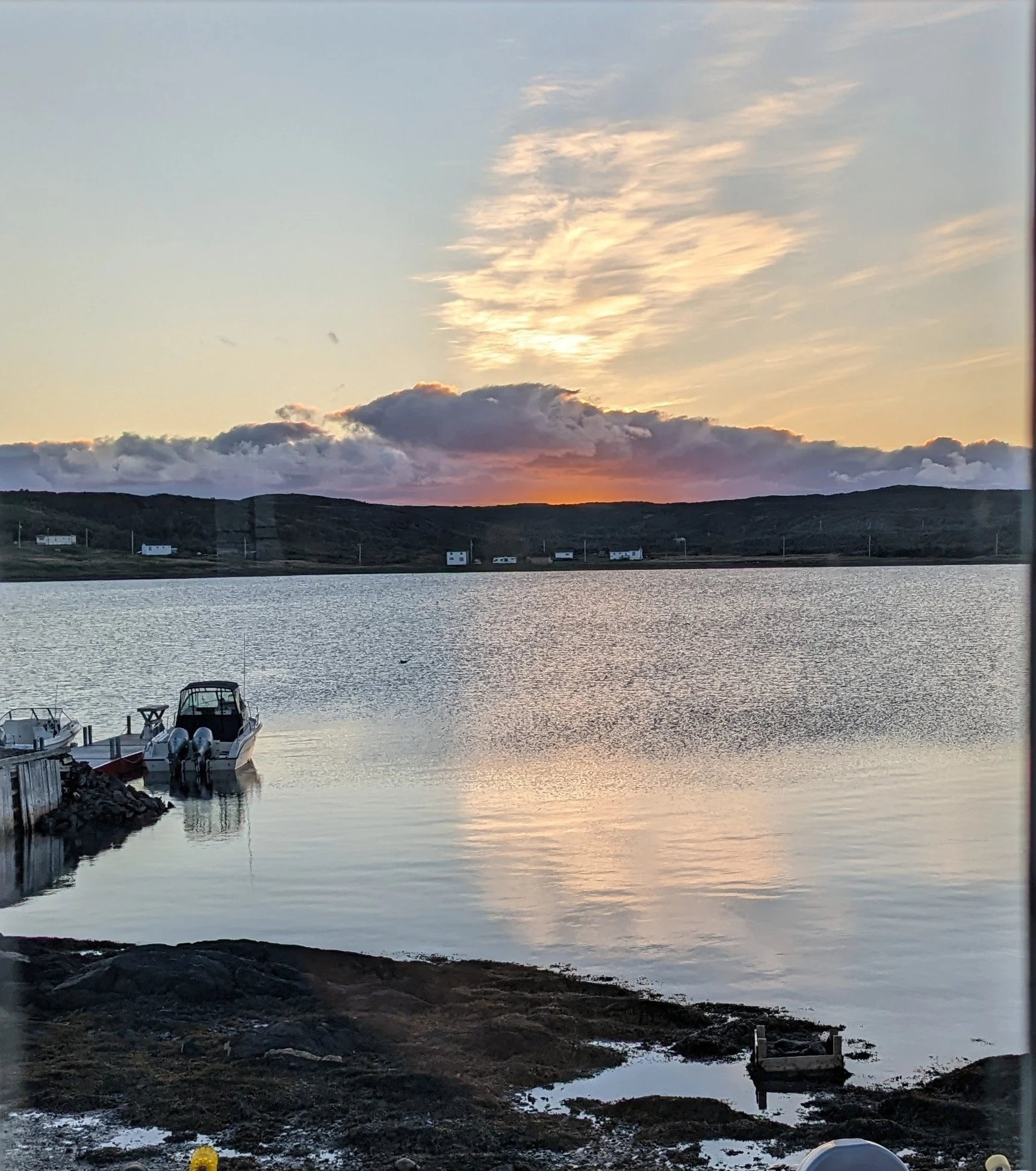 Sunset over a calm body of water with boats docked at a pier, rolling hills in the background, and partly cloudy sky.