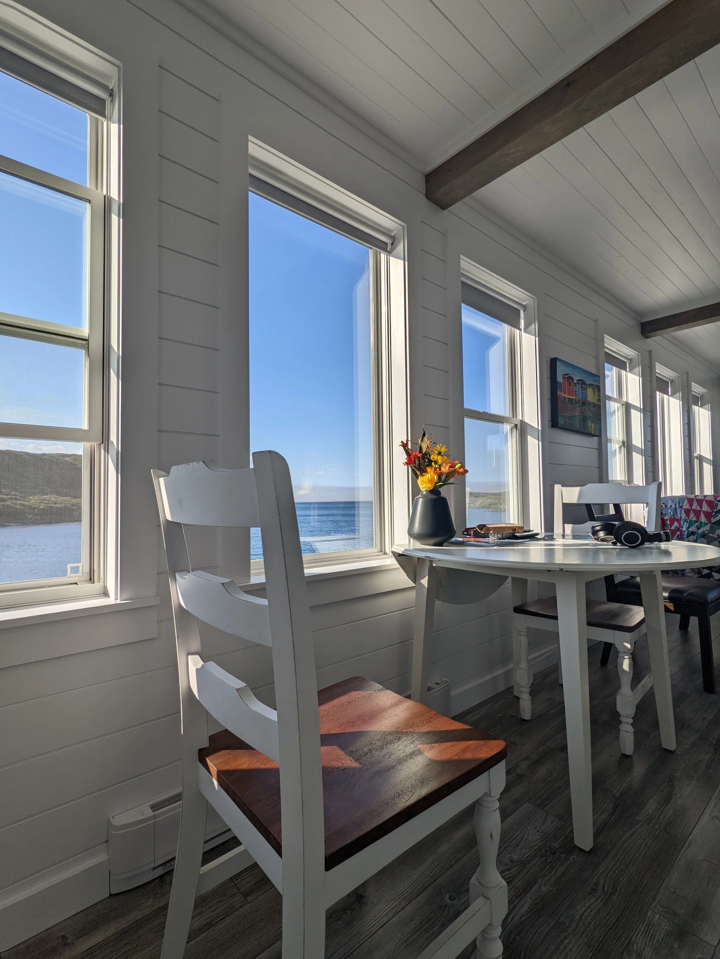 A sunlit coastal room with white wooden walls and large windows looking out over the water with hills on the horizon. Inside, there is a white dining table with two chairs, a black vase with flowers, and some small items, including a pair of headphon
