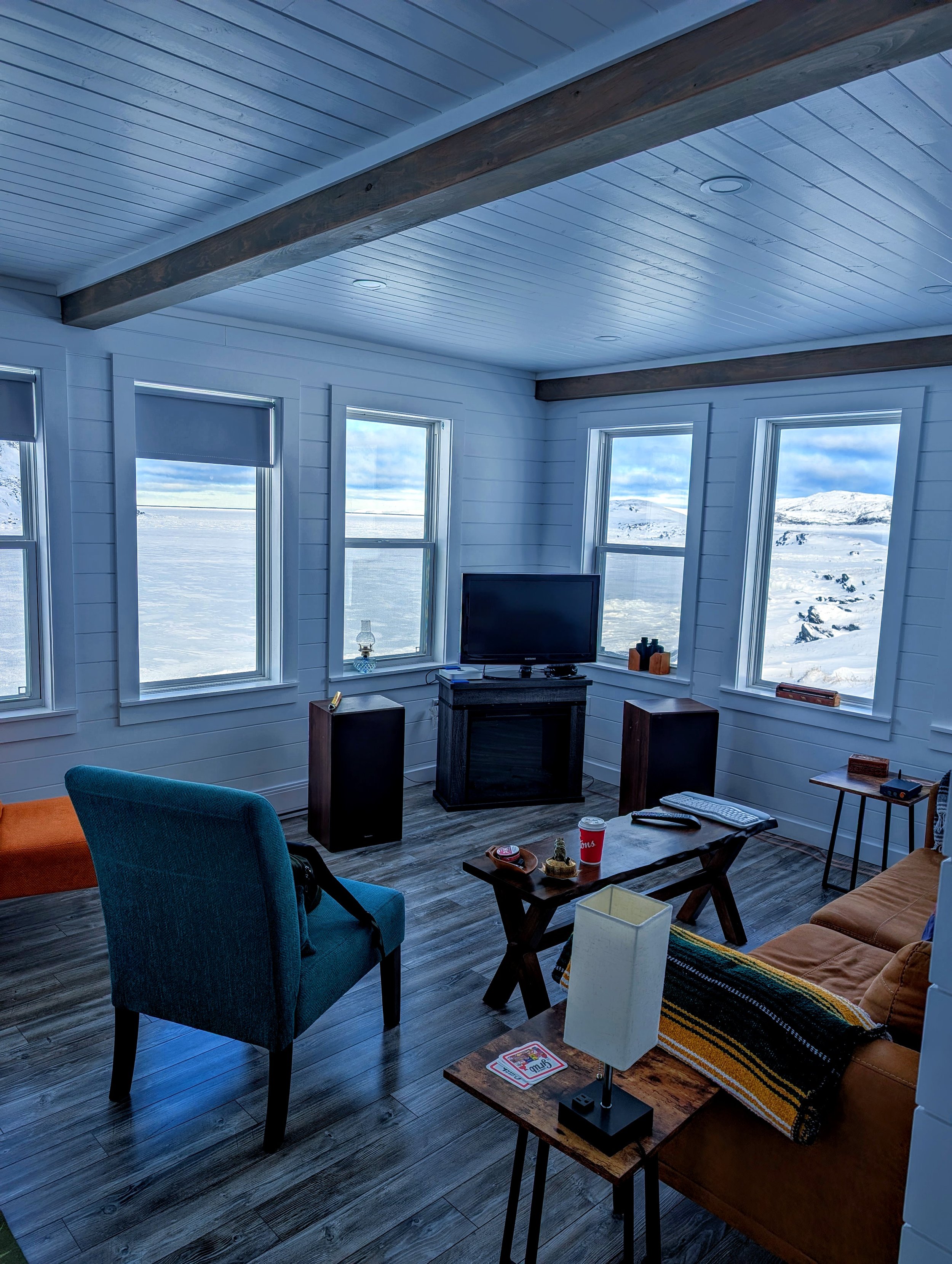 Living room with multiple large windows showing snowy mountains and landscape outside. The room has a wooden ceiling with exposed beams and dark wood flooring. There is a small TV on a stand, chairs, and a coffee table with various items including a 