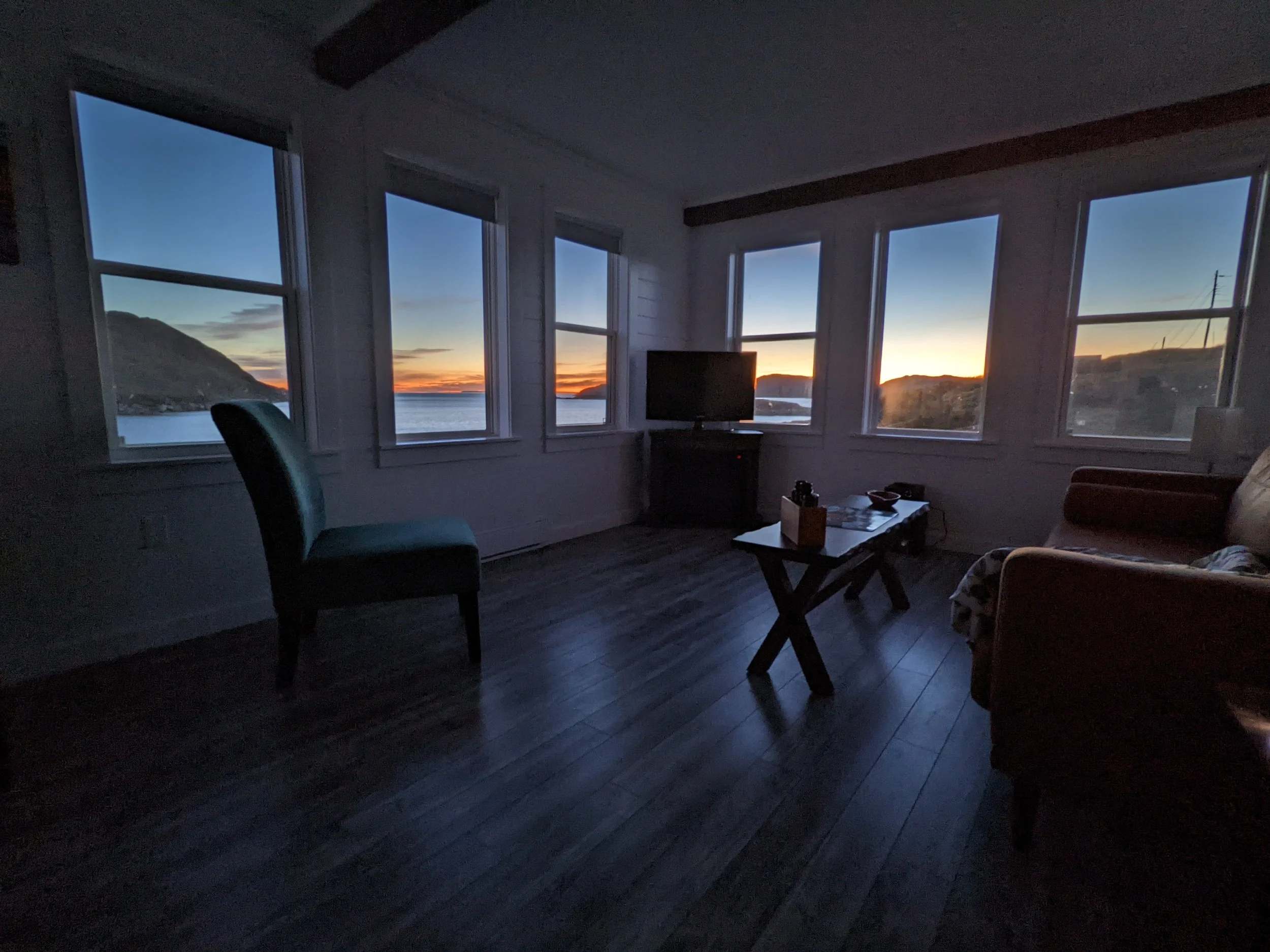 A living room with dark wooden floors and large windows showing a sunset over mountains and water outside.