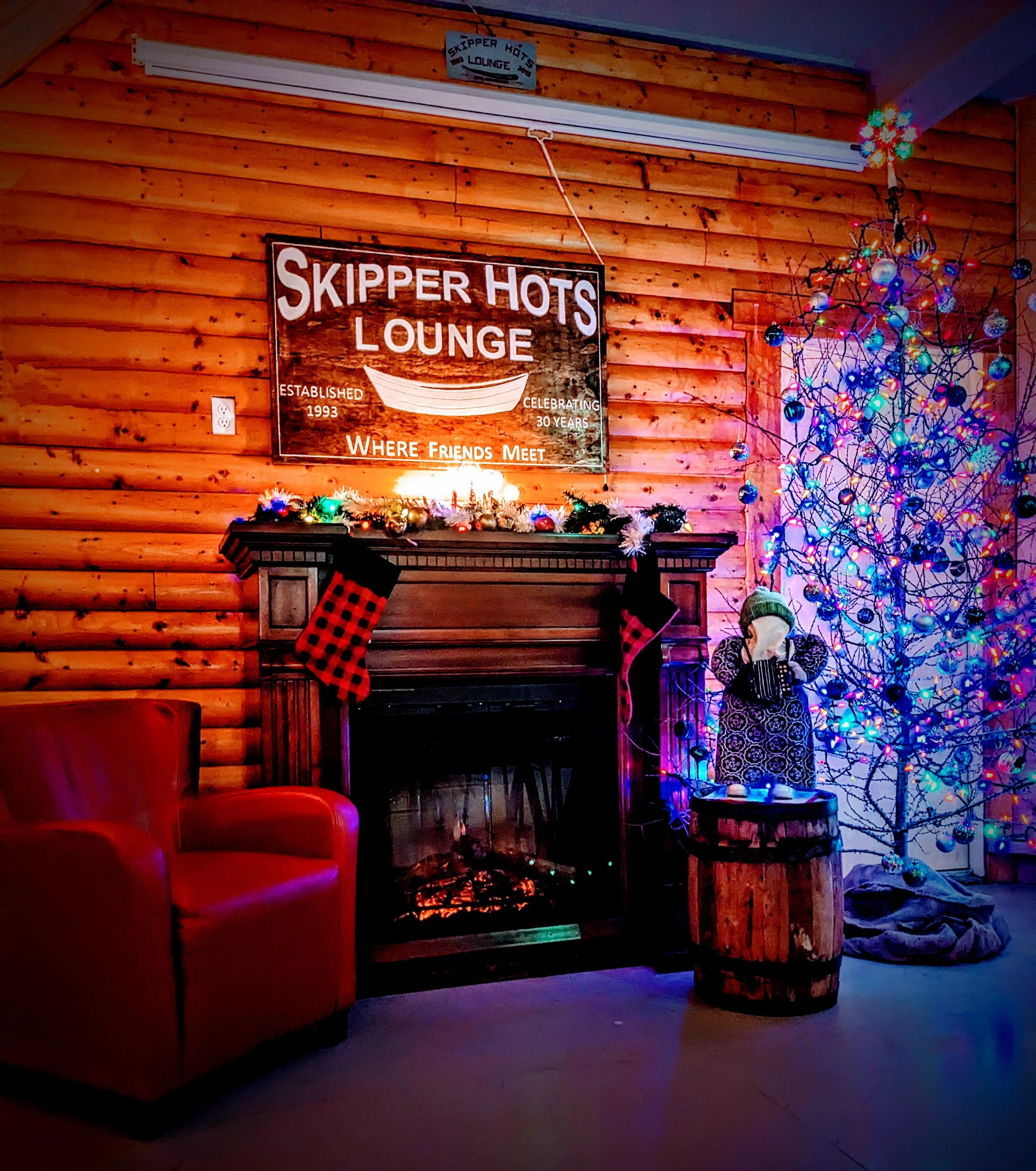 Cozy indoor scene with a neon sign that reads 'Skipper Hots Lounge', a decorated Christmas tree with colorful lights and ornaments, a fireplace with a garland and stockings, and a person in festive attire.
