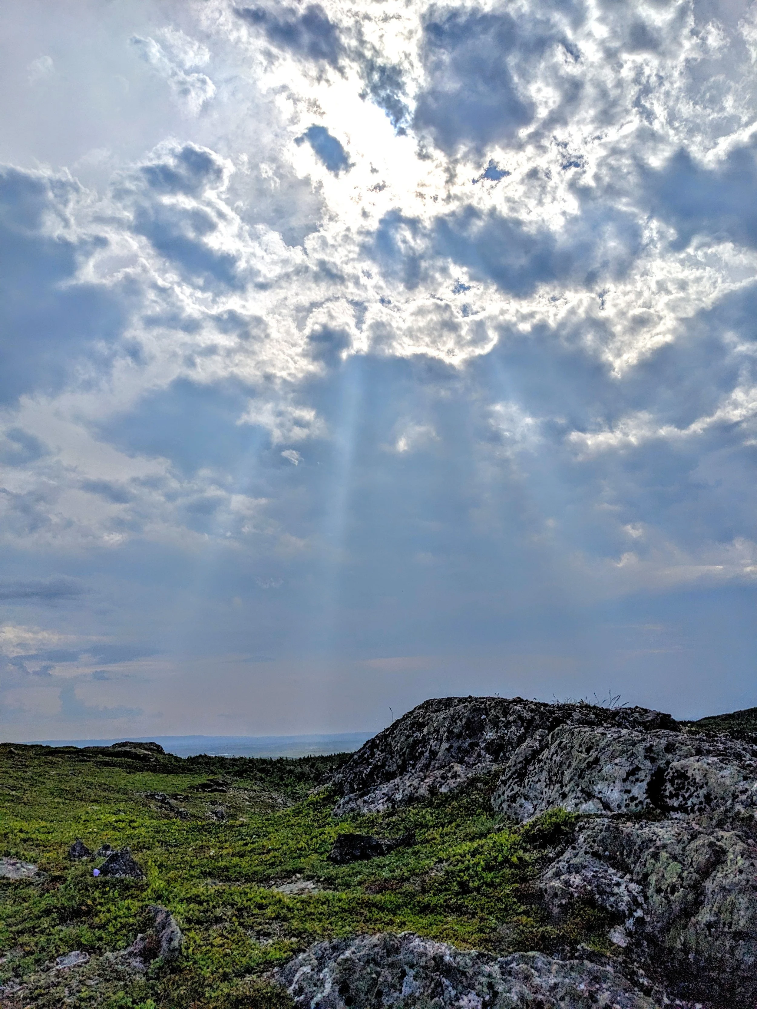 A scenic landscape with large rocks in the foreground, green grass covering the land, and a cloudy sky overhead with sunlight breaking through the clouds.