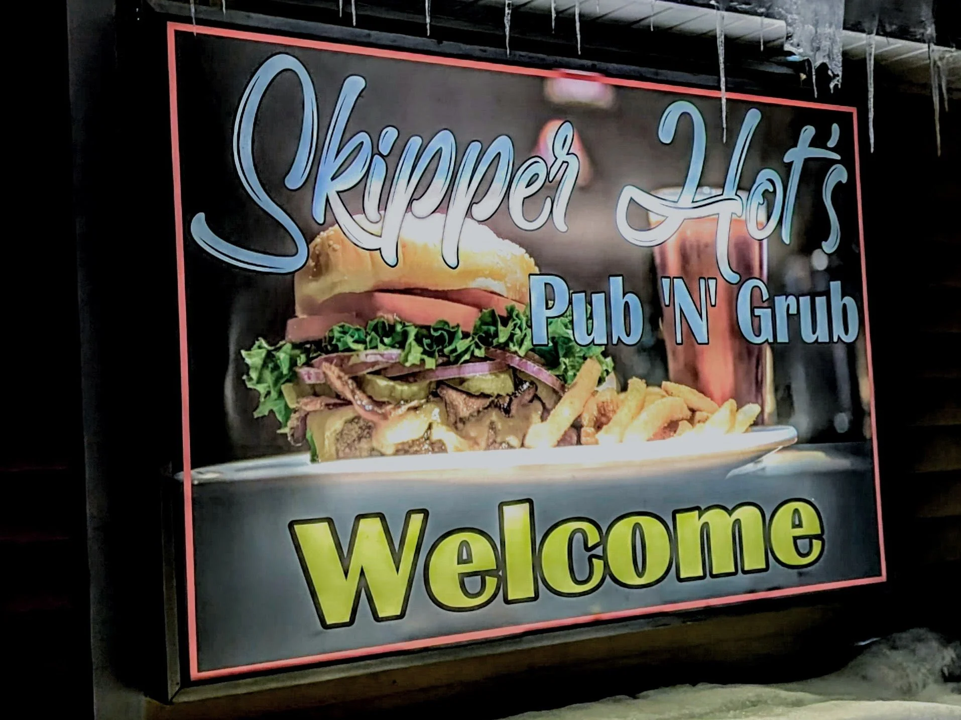 Signboard with the text 'Skipper Hat's Pub 'N' Grub' and a picture of a burger with lettuce, tomato, cheese, bacon, and onion, along with French fries. The sign welcomes customers.