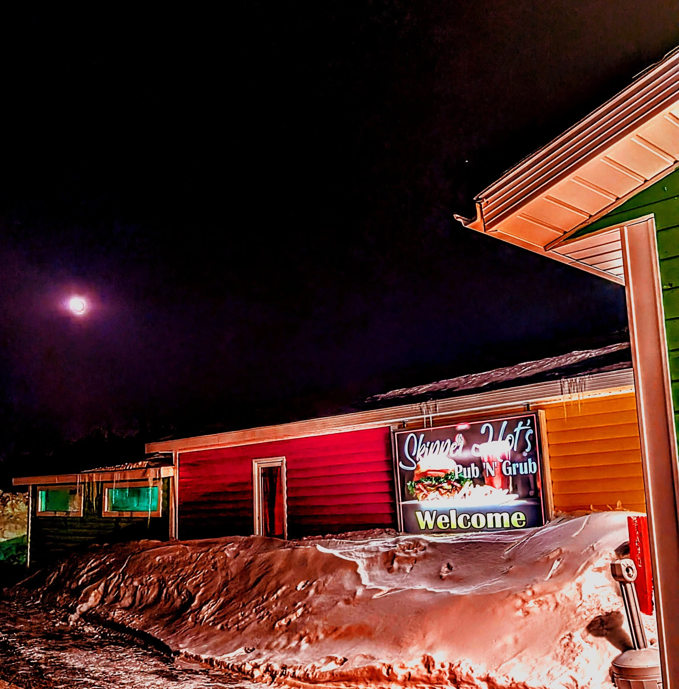 Night scene of a building with a sign that reads 'Skinner's Pub N Grub' and a welcome message. The building has a wooden exterior with a green and red color scheme, and a bright full moon is visible in the dark sky.