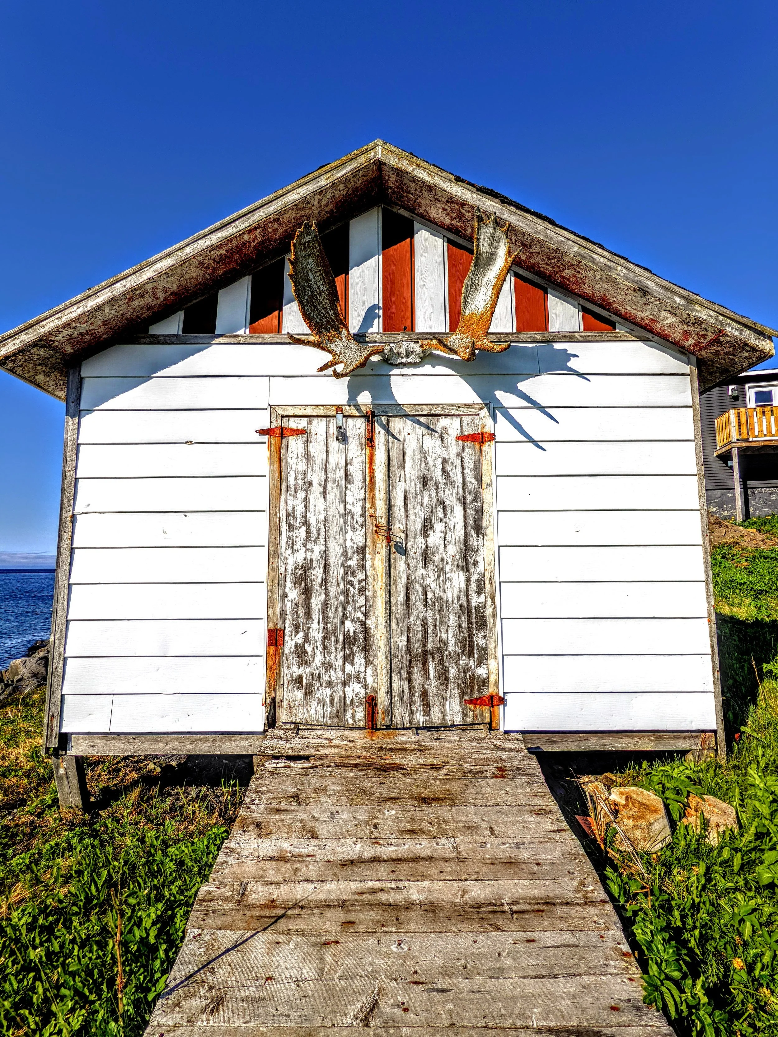 A small white wooden building with a weathered, rusty door and a wooden ramp leading up to it, decorated with a bronze-colored eagle sculpture on the front.