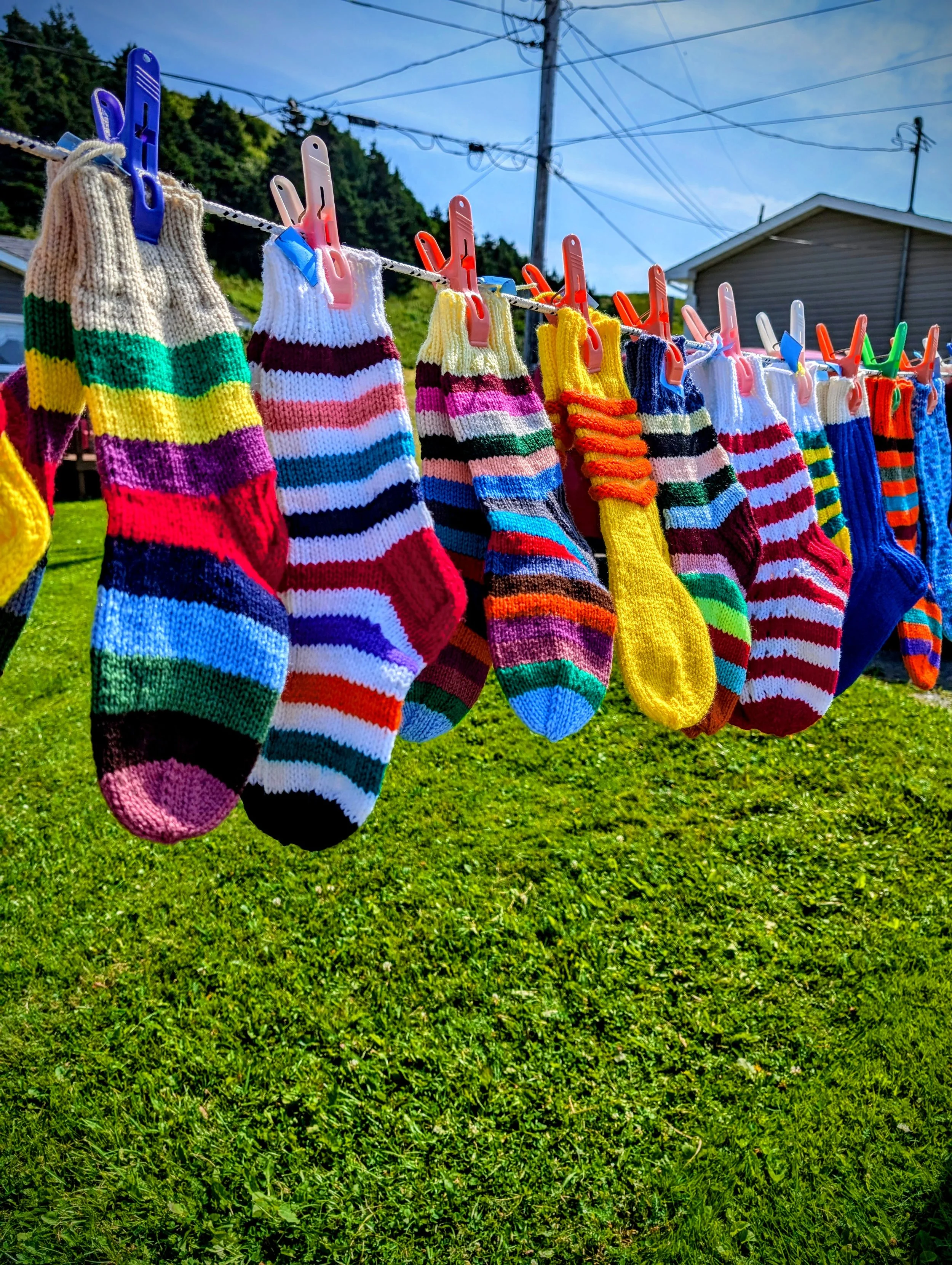 Colorful knitted socks hanging on a clothesline with clothespins in a grassy yard under a blue sky.