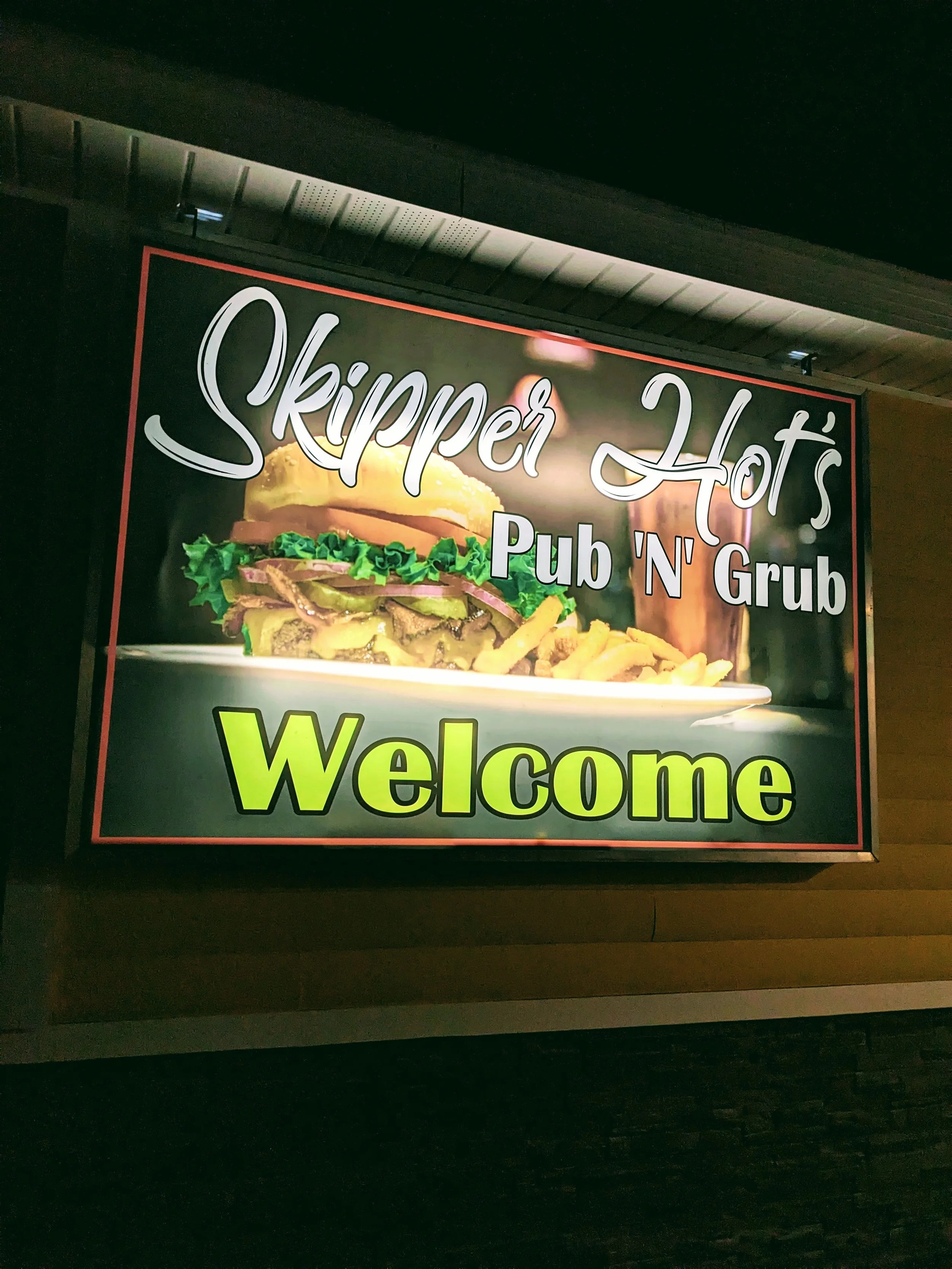 Sign for Skipper Hot's Pub 'N' Grub, featuring an image of a burger with lettuce and fries, and the word 'Welcome' in bright yellow letters.