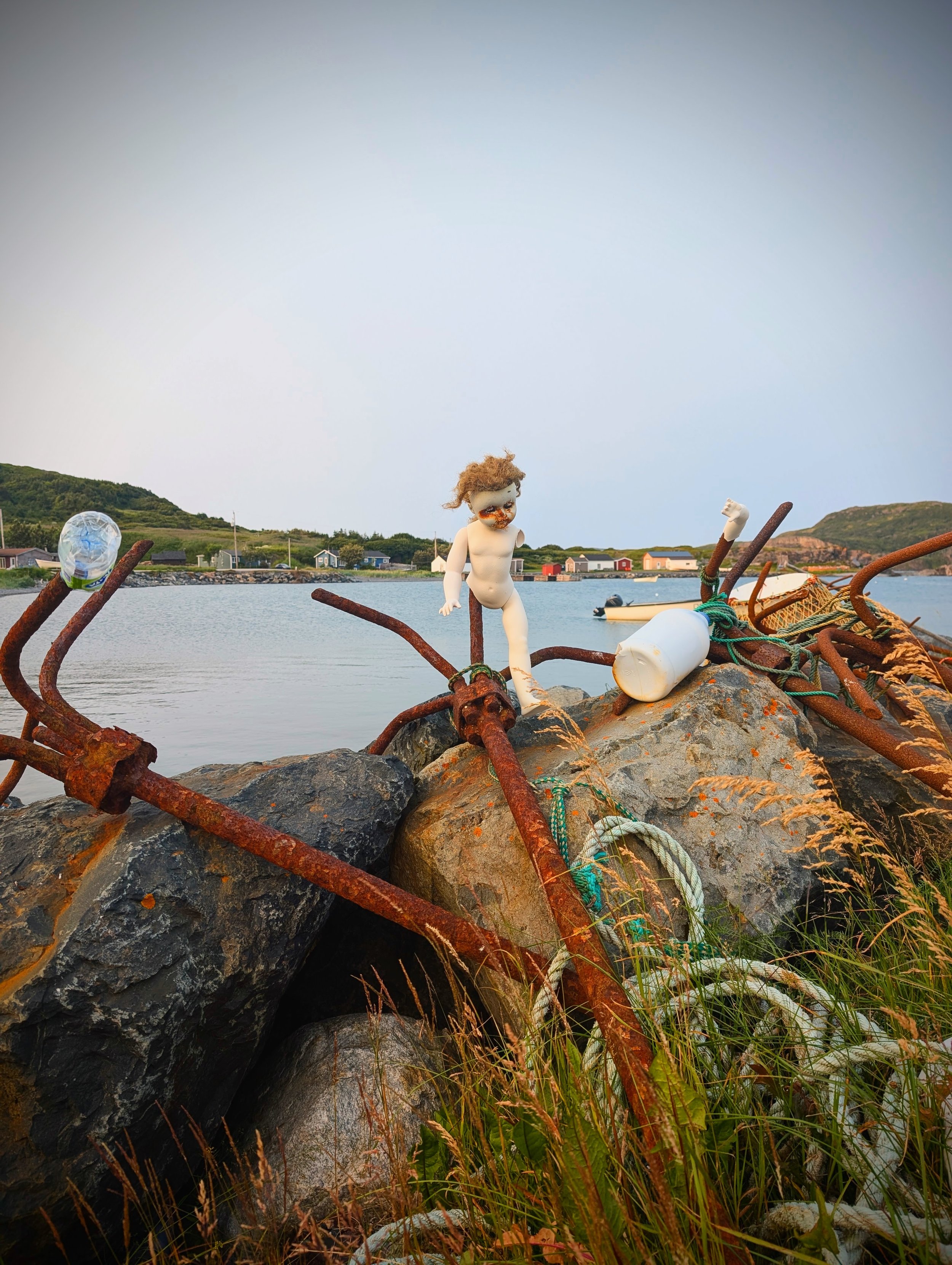 A small doll with curly brown hair, a painted face, and a white body, standing among rusted metal beams, rocks, and tall grass by a body of water with houses and hills in the background.