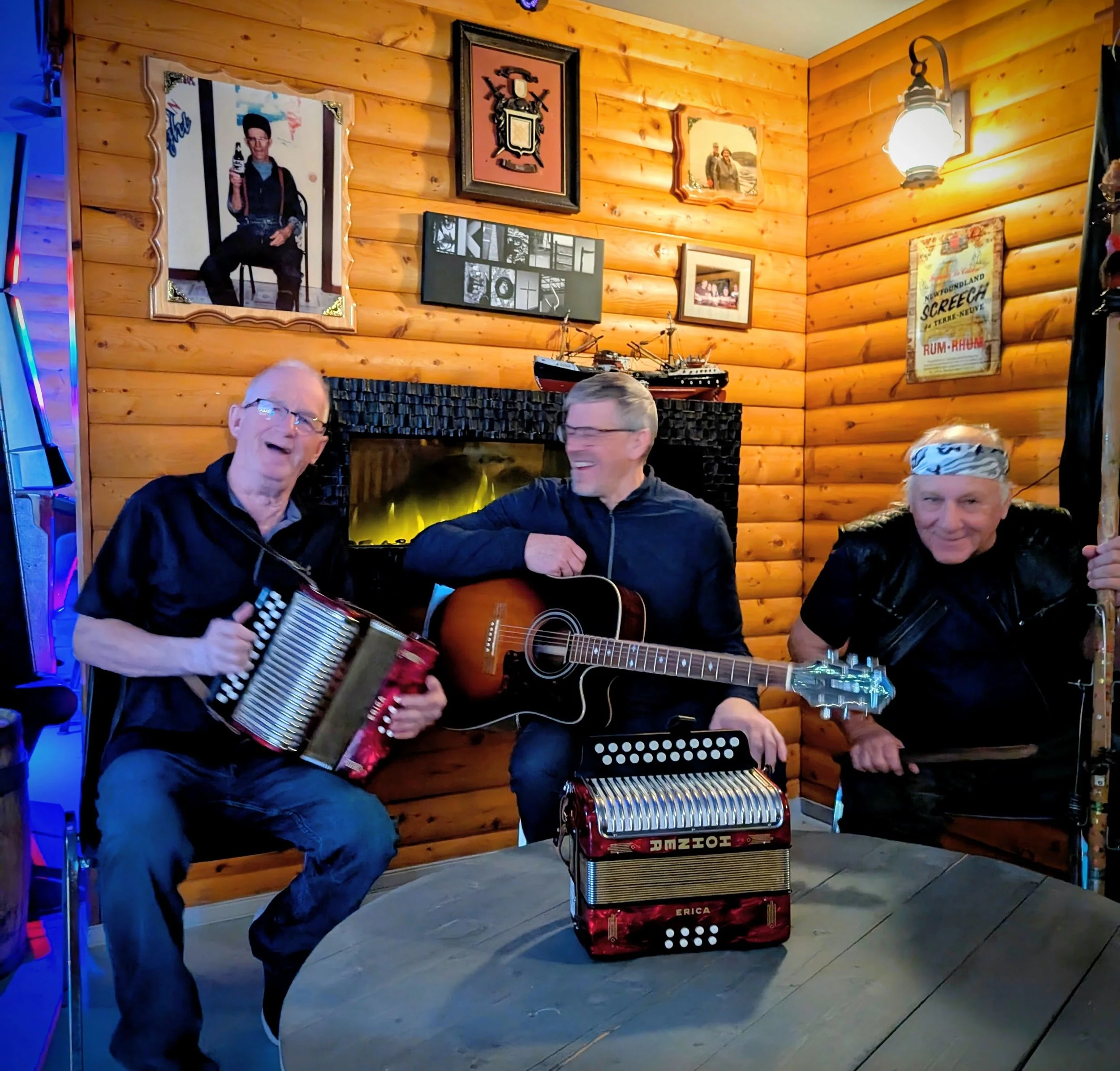 Three men playing musical instruments in a cozy wood-paneled room. One man is playing an accordion, another is playing an acoustic guitar, and the third is playing a keyboard. The background has various framed pictures and posters on the wall, including a large model ship on the shelf, and a lit lantern-style light fixture.