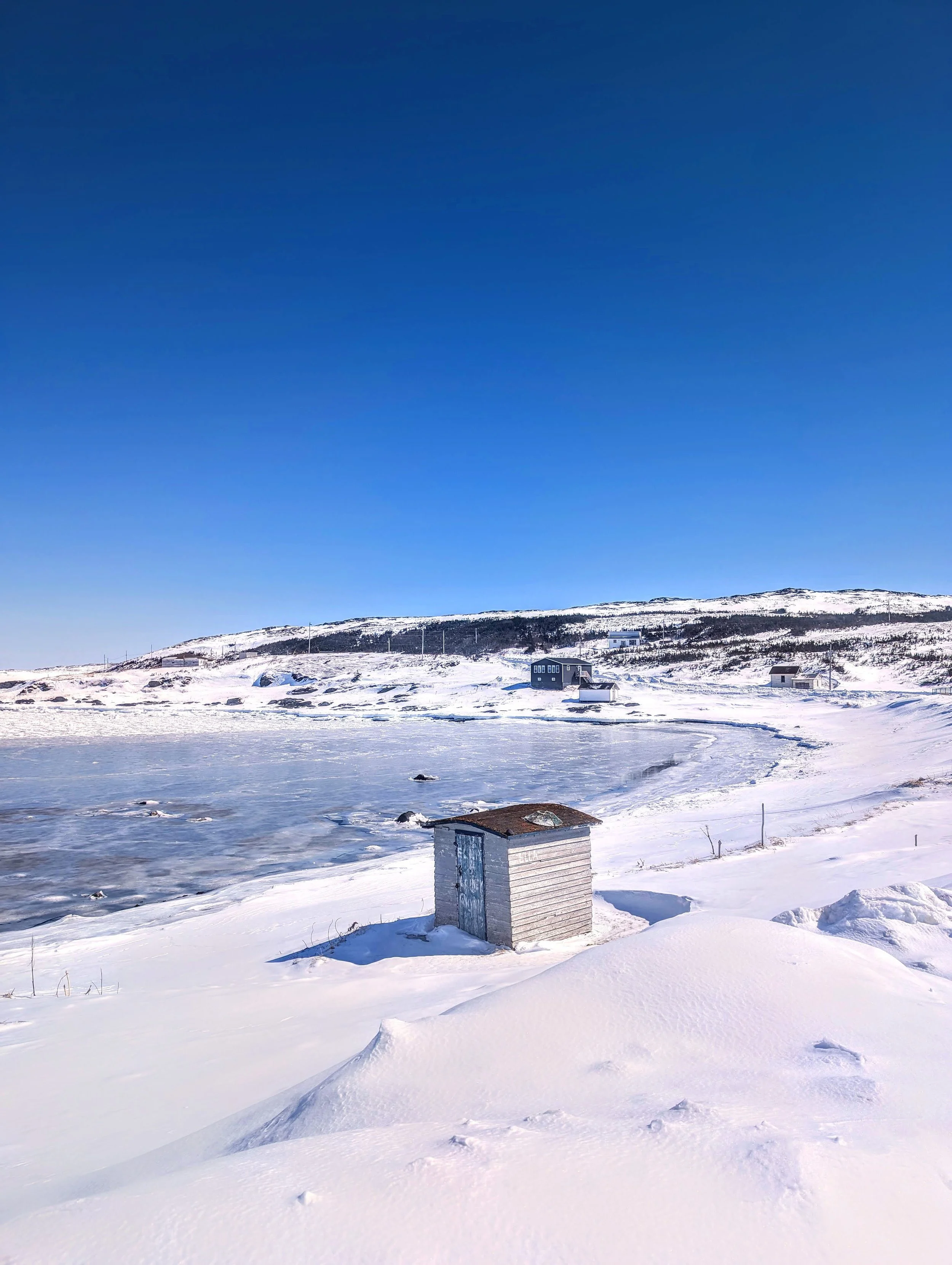 Snow-covered landscape with a frozen body of water and small buildings, under a clear blue sky.