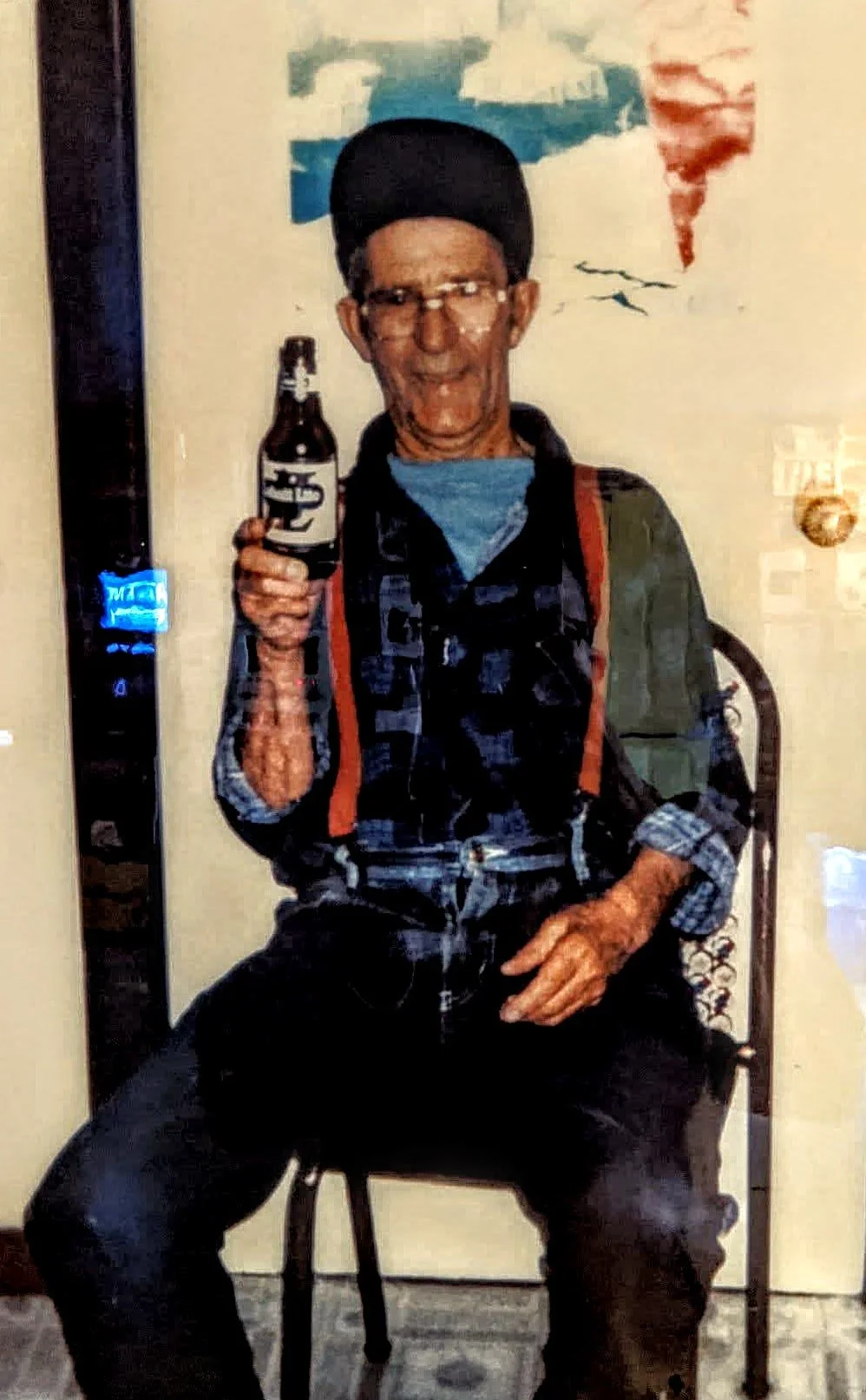 An elderly man with glasses, wearing a black beanie and a dark flannel shirt, holding a bottle in his right hand, sitting on a chair with a green backpack on his back, in front of a painted wall with clouds.