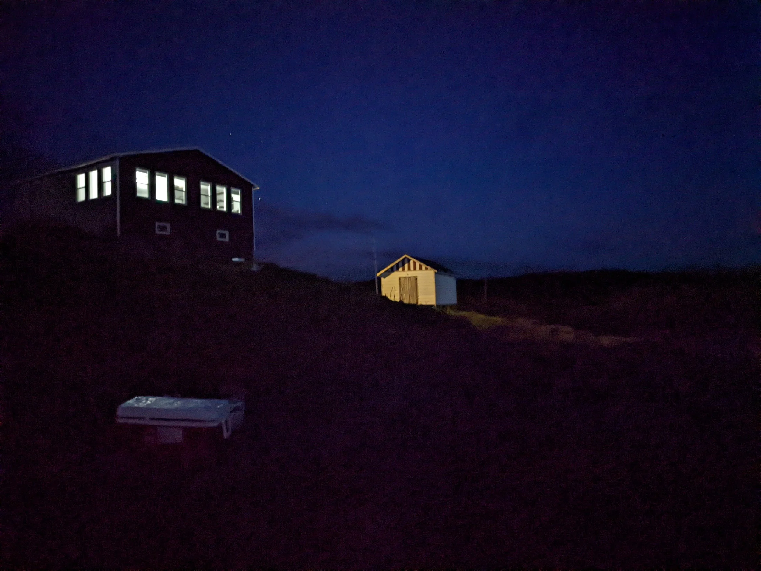 A dark nighttime scene showing a house with lit windows, a small shed, and a white object in the foreground, all set against a dark sky.