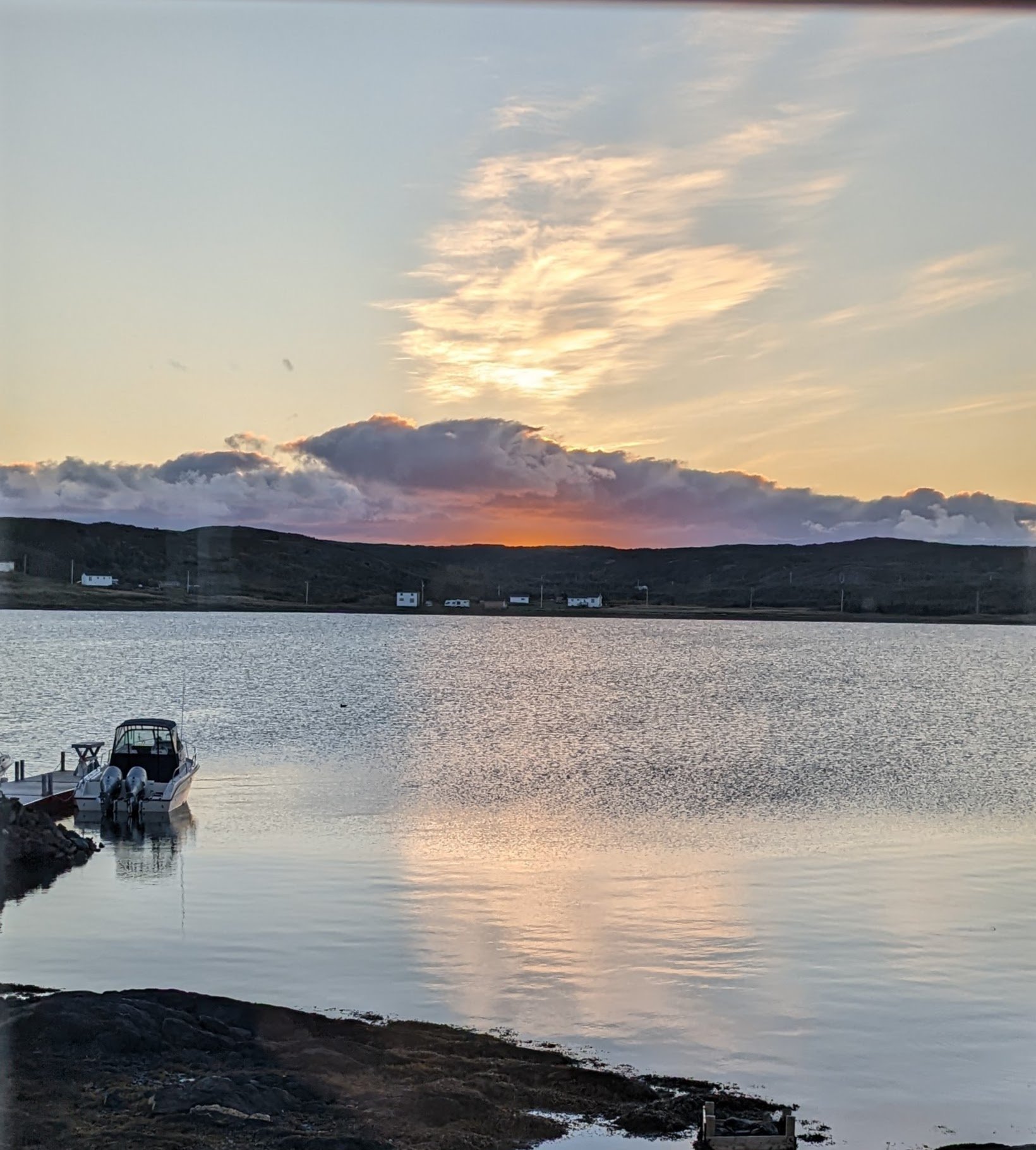 Sunset over a calm body of water with hills in the background, two small white houses along the shore, and boats docked near the rocks in the foreground.