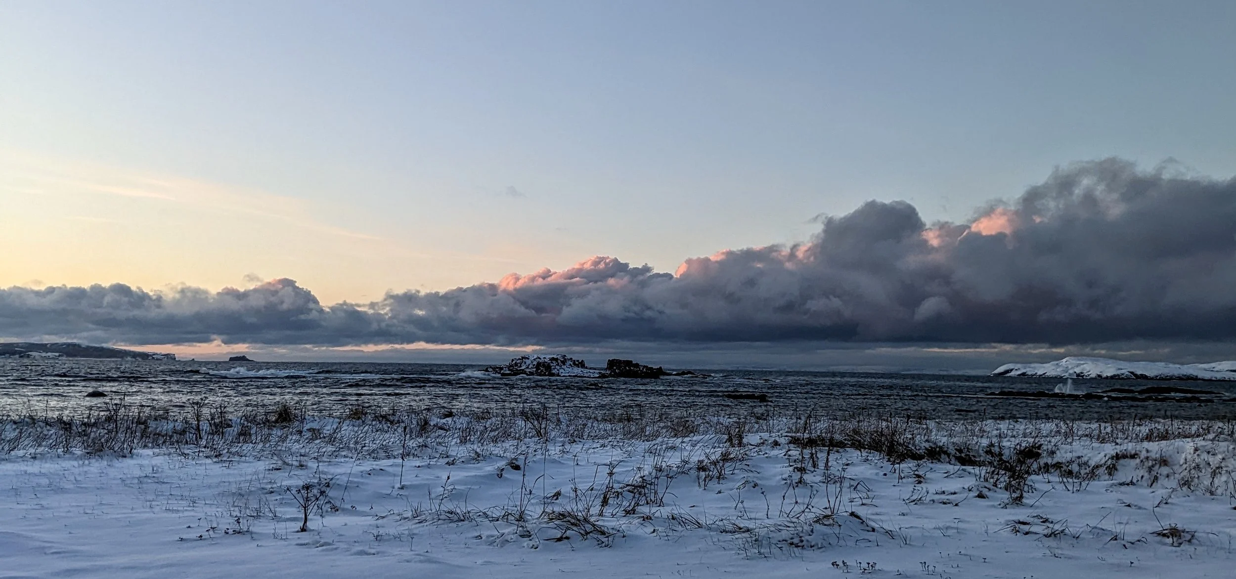 A winter landscape showing snow-covered ground, dark clouds in the sky with pink highlights, and a distant view of coastline with snow-capped cliffs.