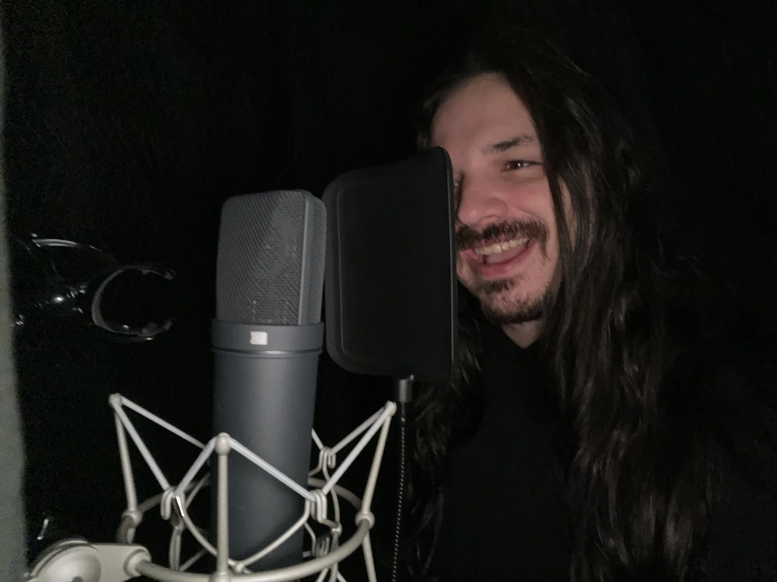 A man with long dark hair and a beard smiling behind a microphone in a recording studio.