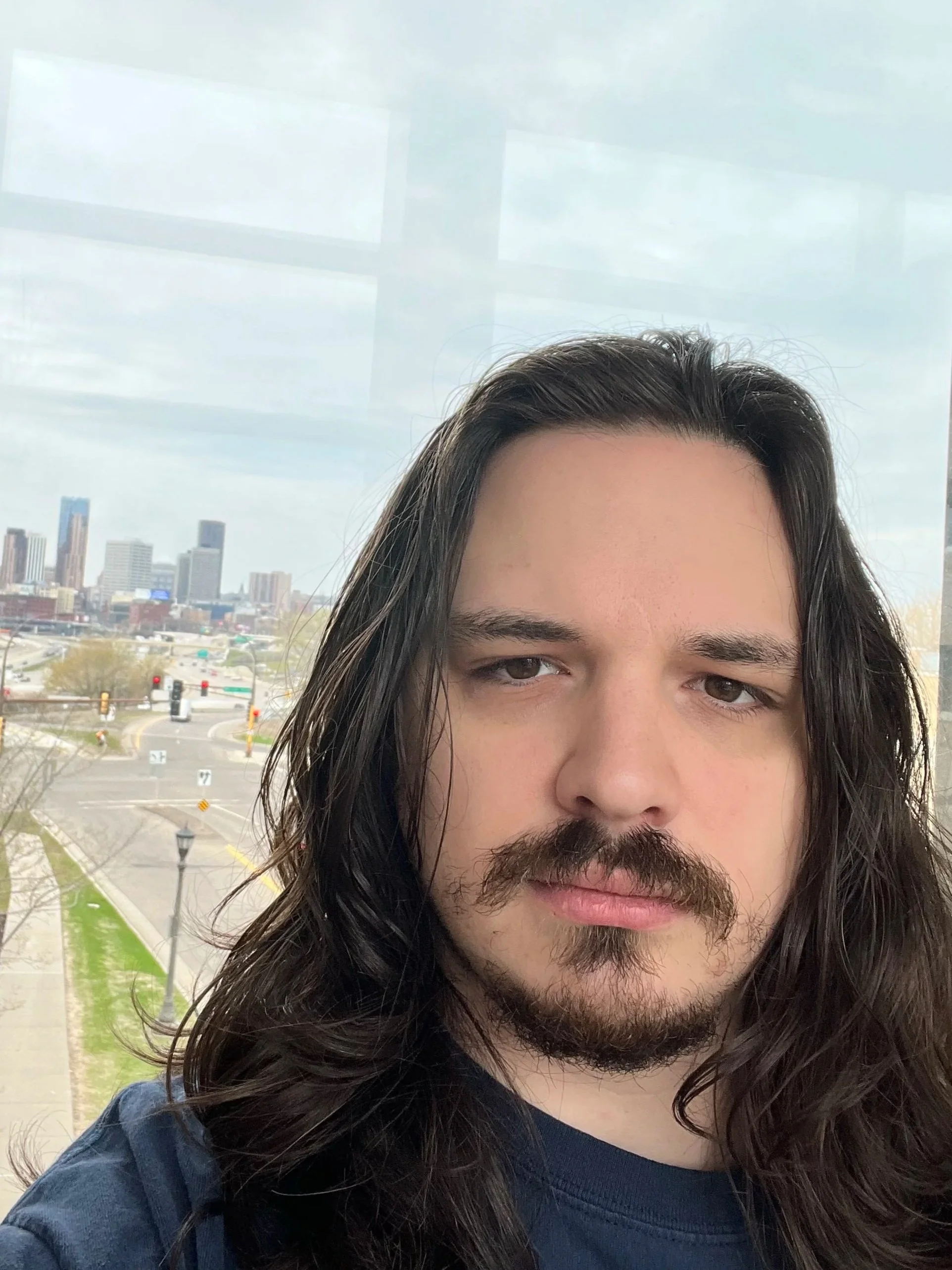 A man with long dark hair and a beard taking a selfie indoors, with a cityscape visible through a large window behind him.