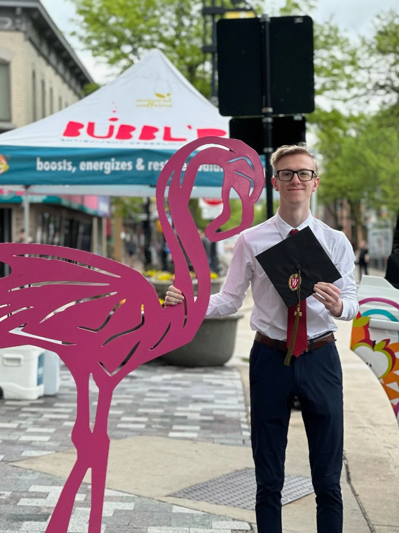 A young man dressed in a white shirt, red tie, and glasses holds a black graduation cap and stands behind a pink flamingo cutout during a graduation celebration outdoors. In the background, there are tents, trees, and a street scene.