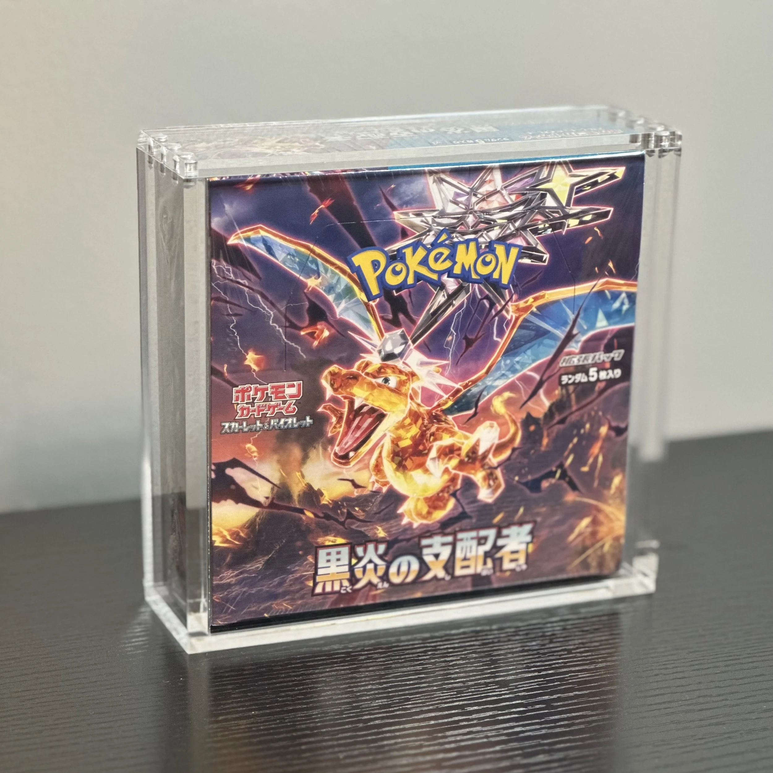 Close-up of a Pokémon trading card game booster box inside a clear acrylic case, featuring artwork of a Pokémon surrounded by lightning bolts on the packaging.