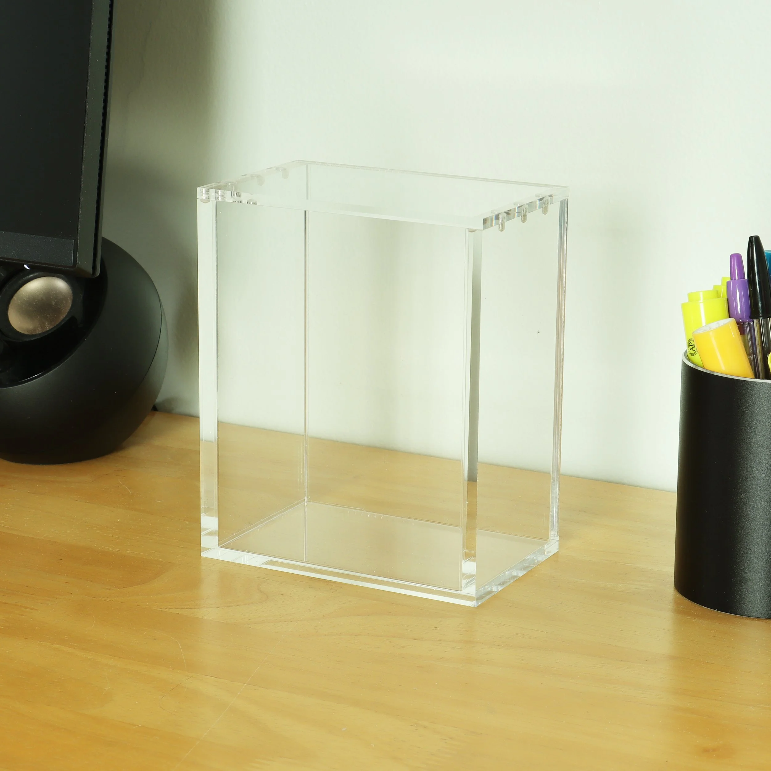 Clear acrylic display stand on a wooden surface with various colored highlighters and a black speaker in the background.