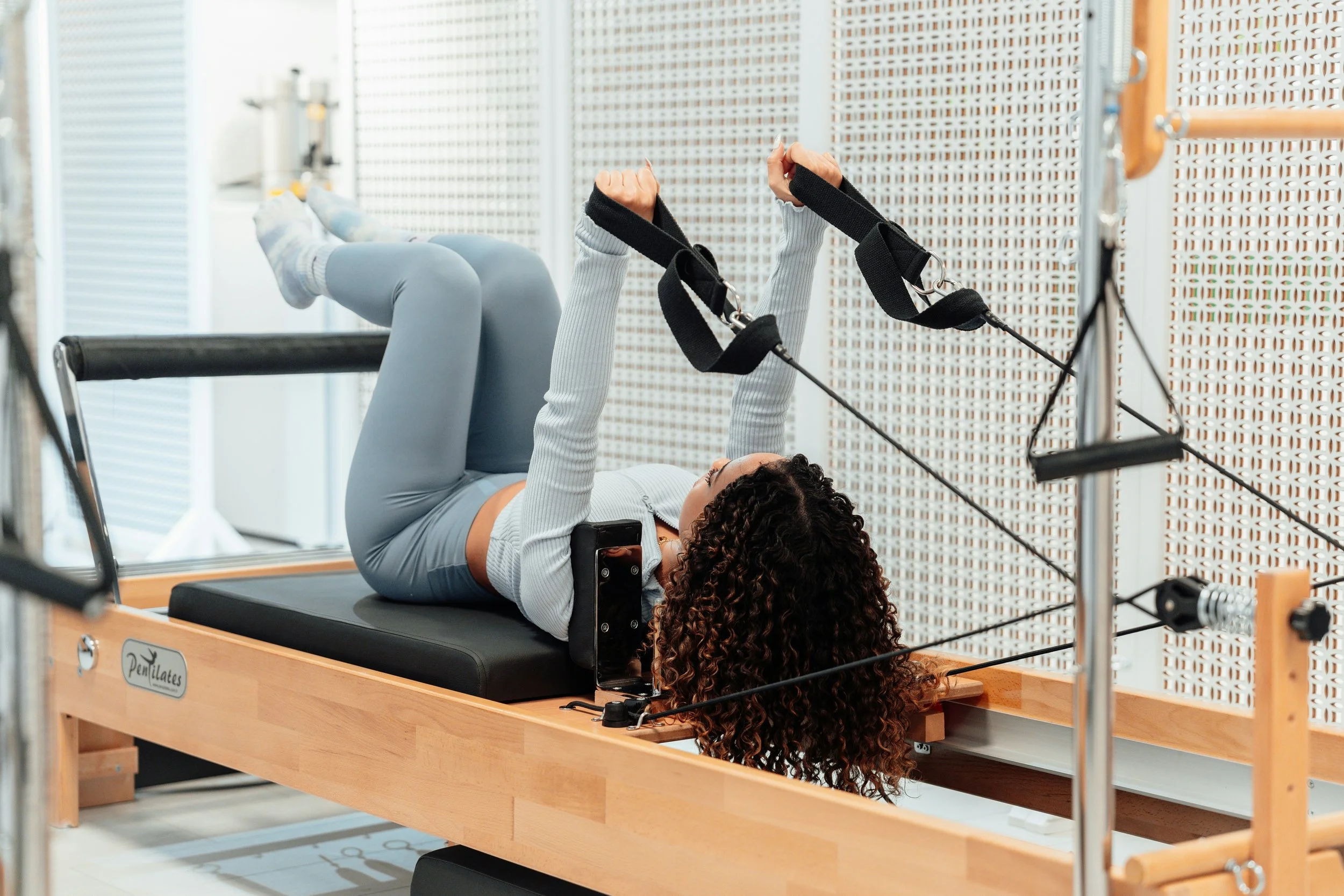 A woman lying on her back on a reformer Pilates machine, holding resistance straps with both hands while exercising, with curly hair and a smartphone placed on her waist.