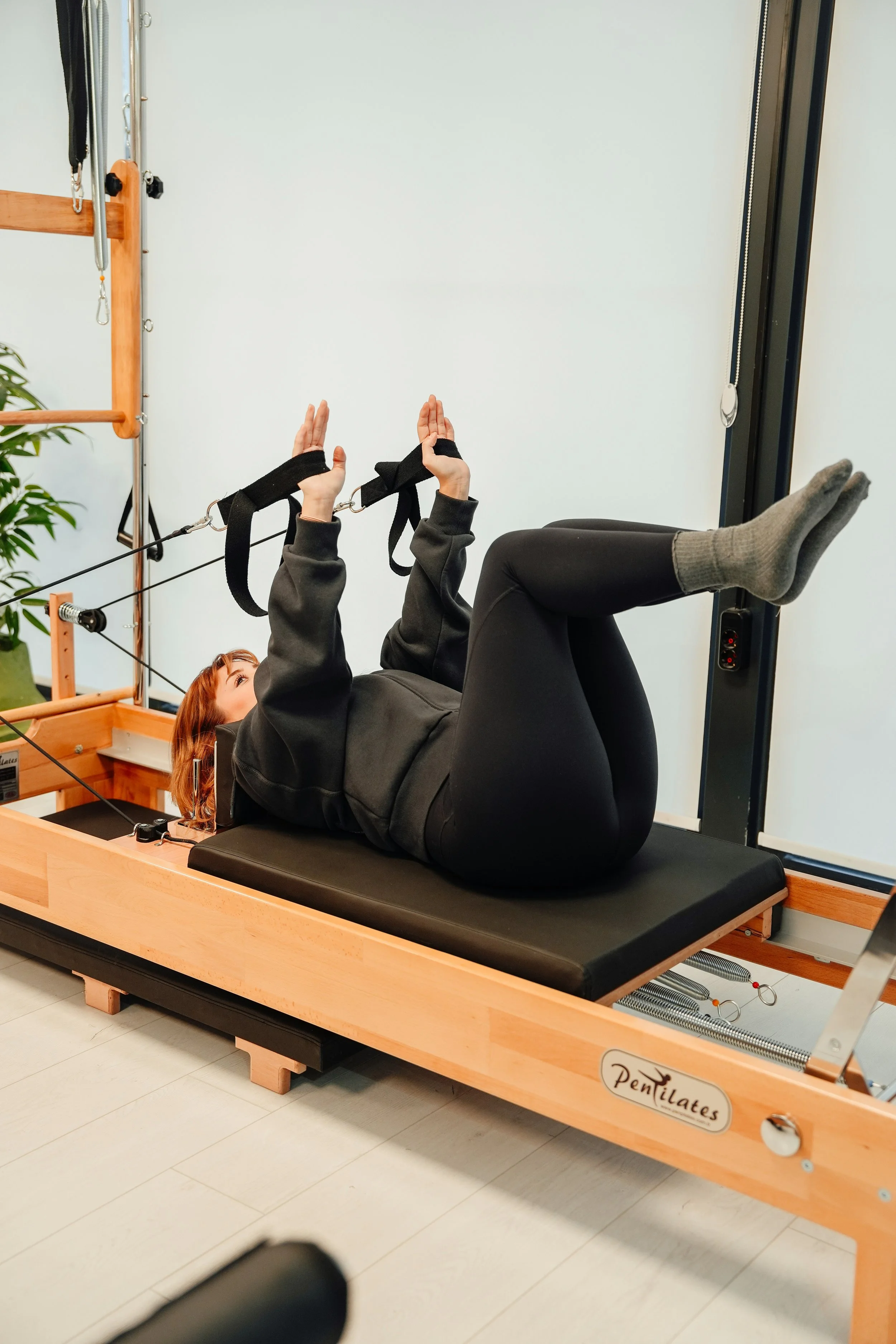 A person with red hair lying on a Pilates reformer machine, legs bent, holding straps with hands, in a workout studio.