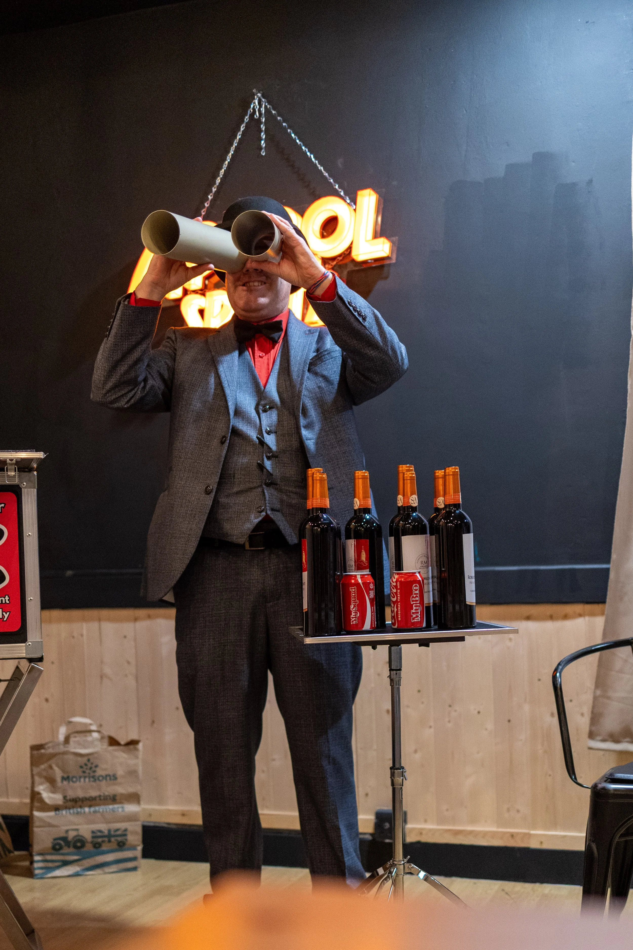 A man in a gray suit and red shirt is looking through binoculars in front of a bar counter with several bottles of wine and cans of soda. Behind him, there is a black wall with a neon sign and a chain hanging from the ceiling.