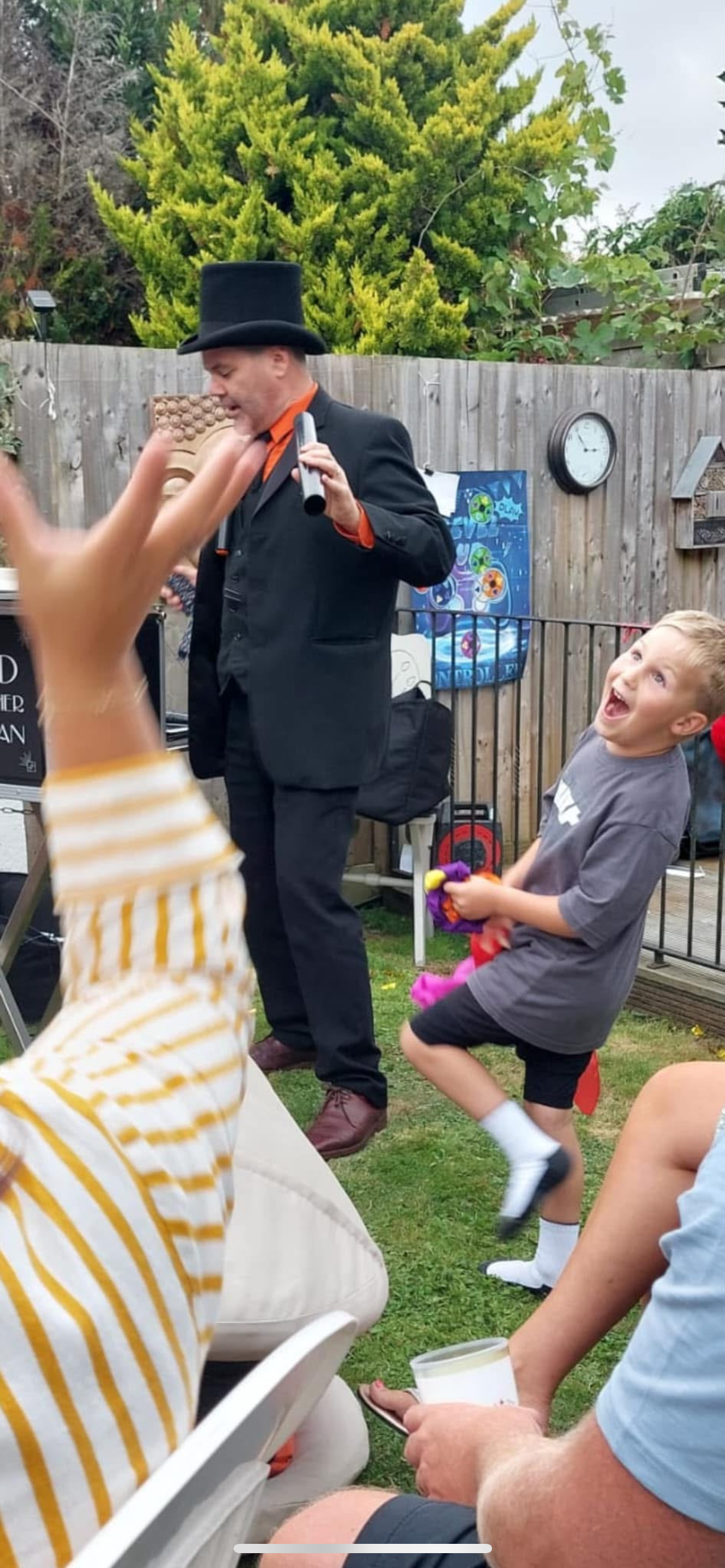 A man dressed as a magician with a black top hat and suit entertains children at an outdoor backyard party. He is holding a microphone and a magic wand, while a boy in a gray T-shirt and black shorts is excited, holding colorful balloons. Other children and adults are seated and watching, with a wooden fence, clocks, and posters in the background.