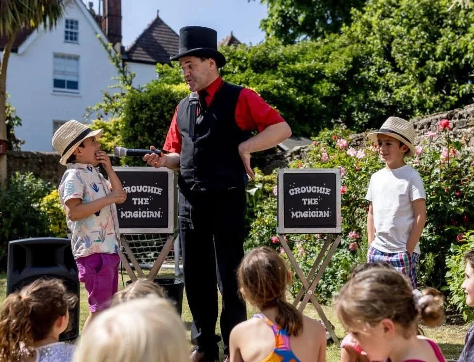 A magician in a black top hat performing for children outdoors, with two boys on either side and an audience in front, during daytime in a garden with trees and flowers.