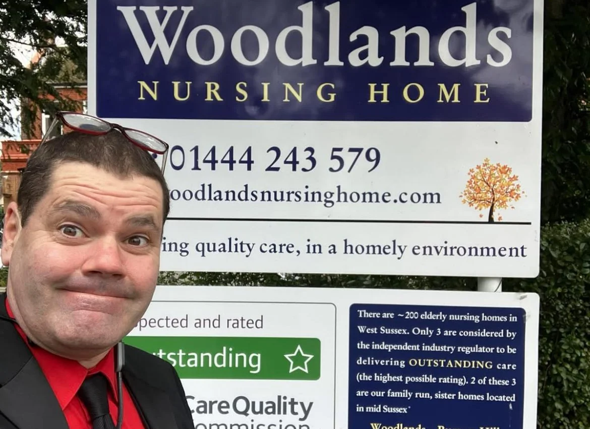 A man taking a selfie in front of a sign for Woodlands Nursing Home, which provides elderly care in West Sussex. The sign includes contact information, a website, and a note about the home's high standard of care.