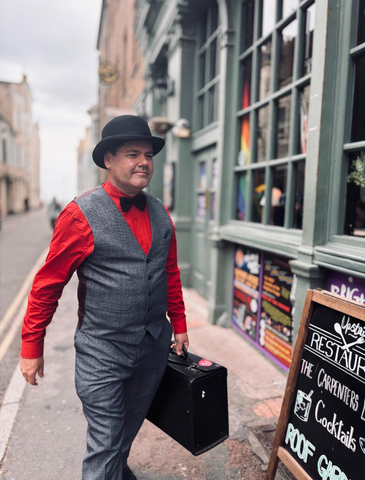 A man dressed in a red shirt, grey vest, black bow tie, and black hat standing on a city sidewalk holding a black briefcase, with a storefront and street scene in the background.